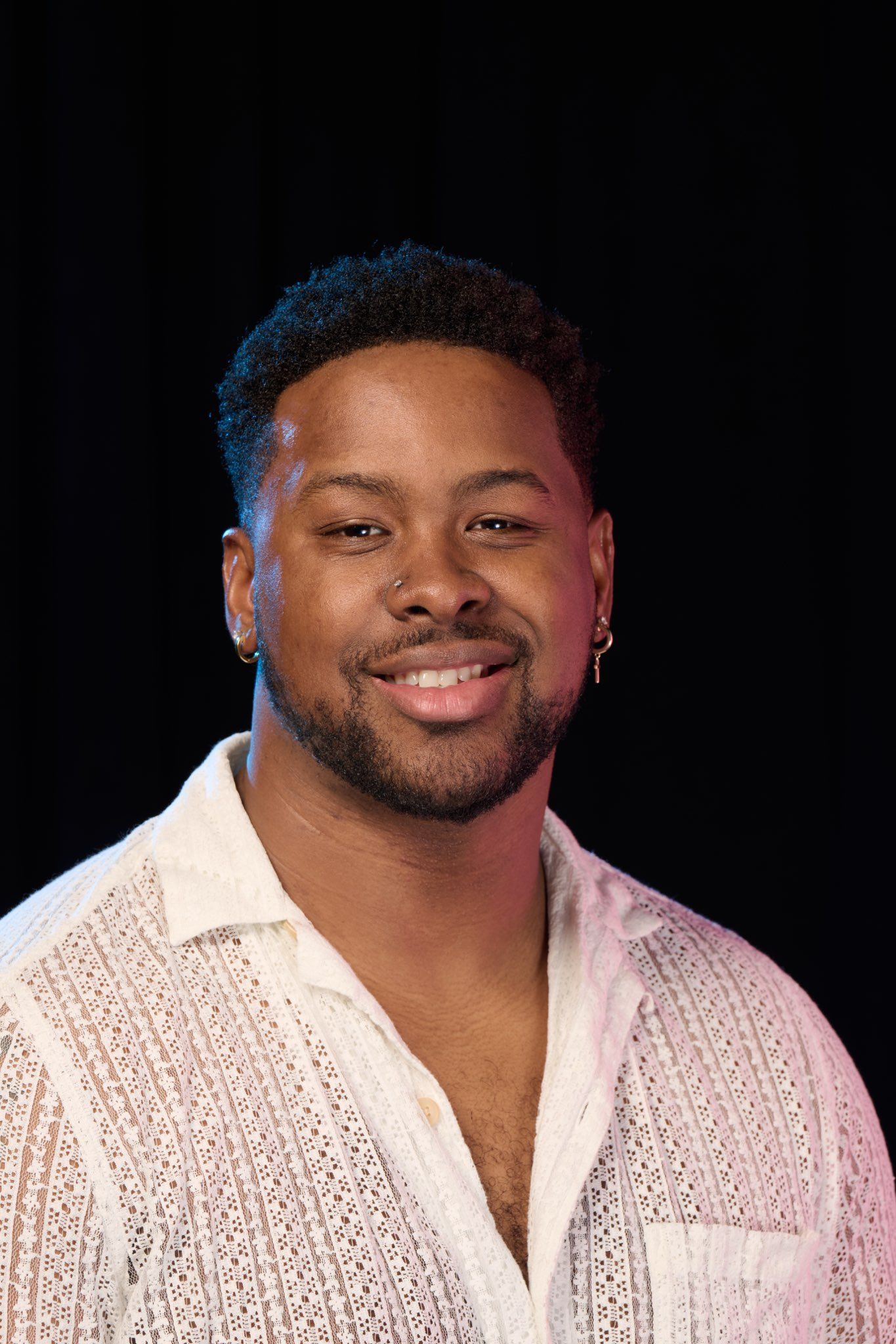 Portrait of a smiling young man with short hair wearing a white lace shirt against a black background.