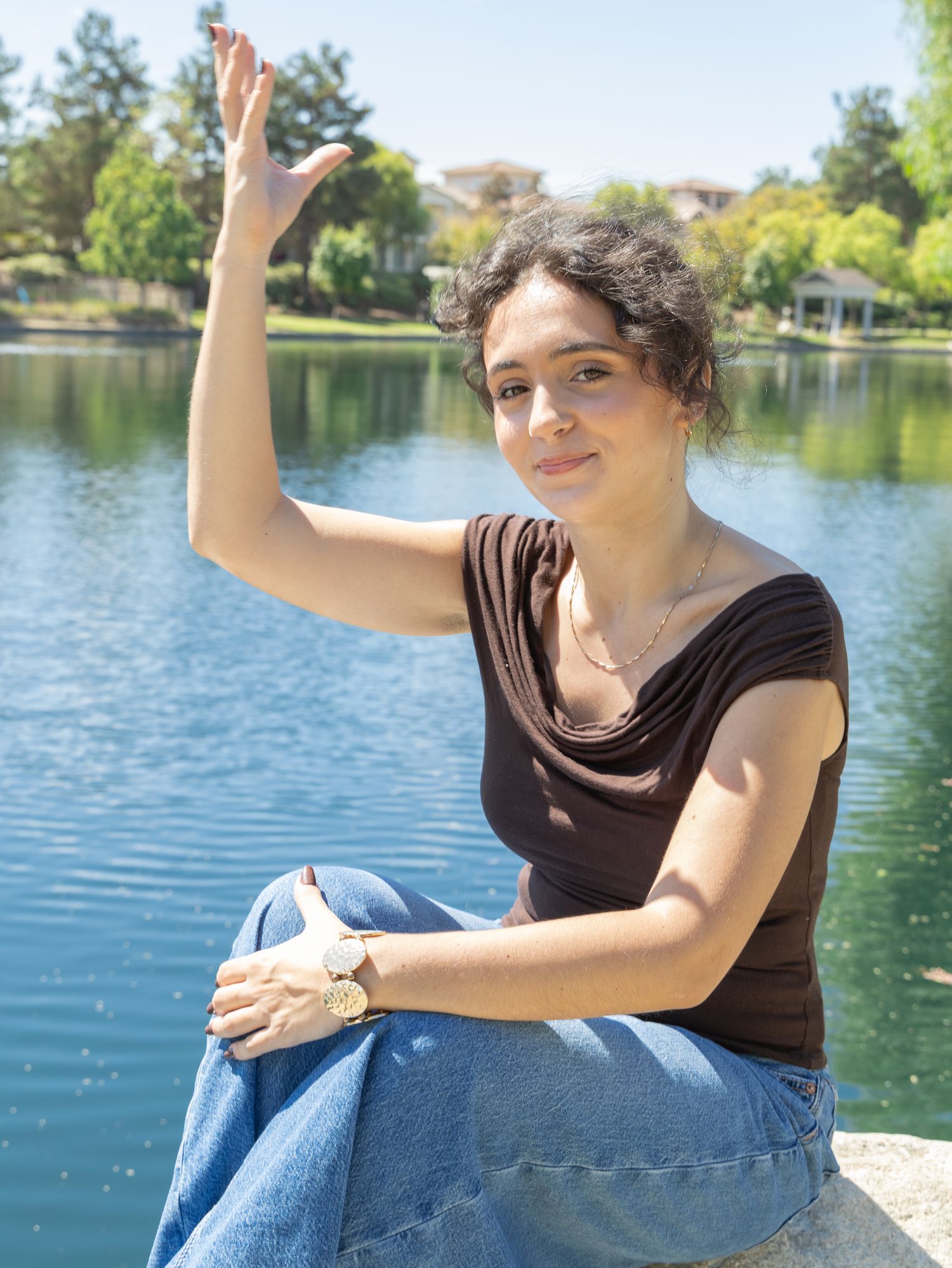 Woman waving hand by a scenic lake, wearing brown top and jeans, sitting outdoors on a sunny day.