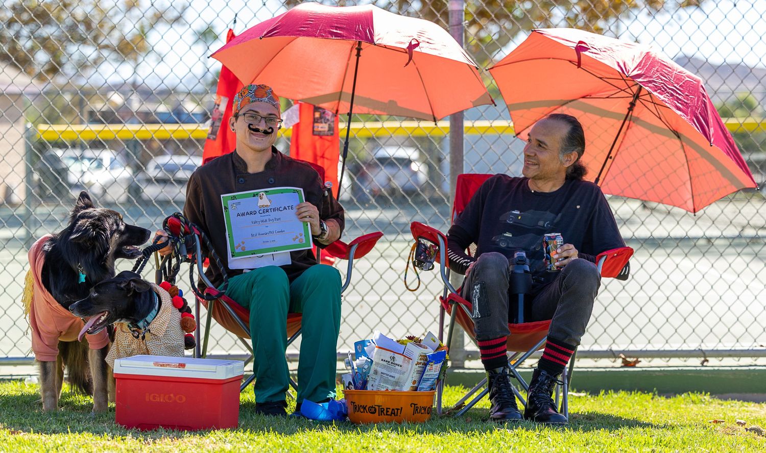 Two spectators sit in chairs under red umbrellas at an outdoor sports field with a dog and cooler nearby.