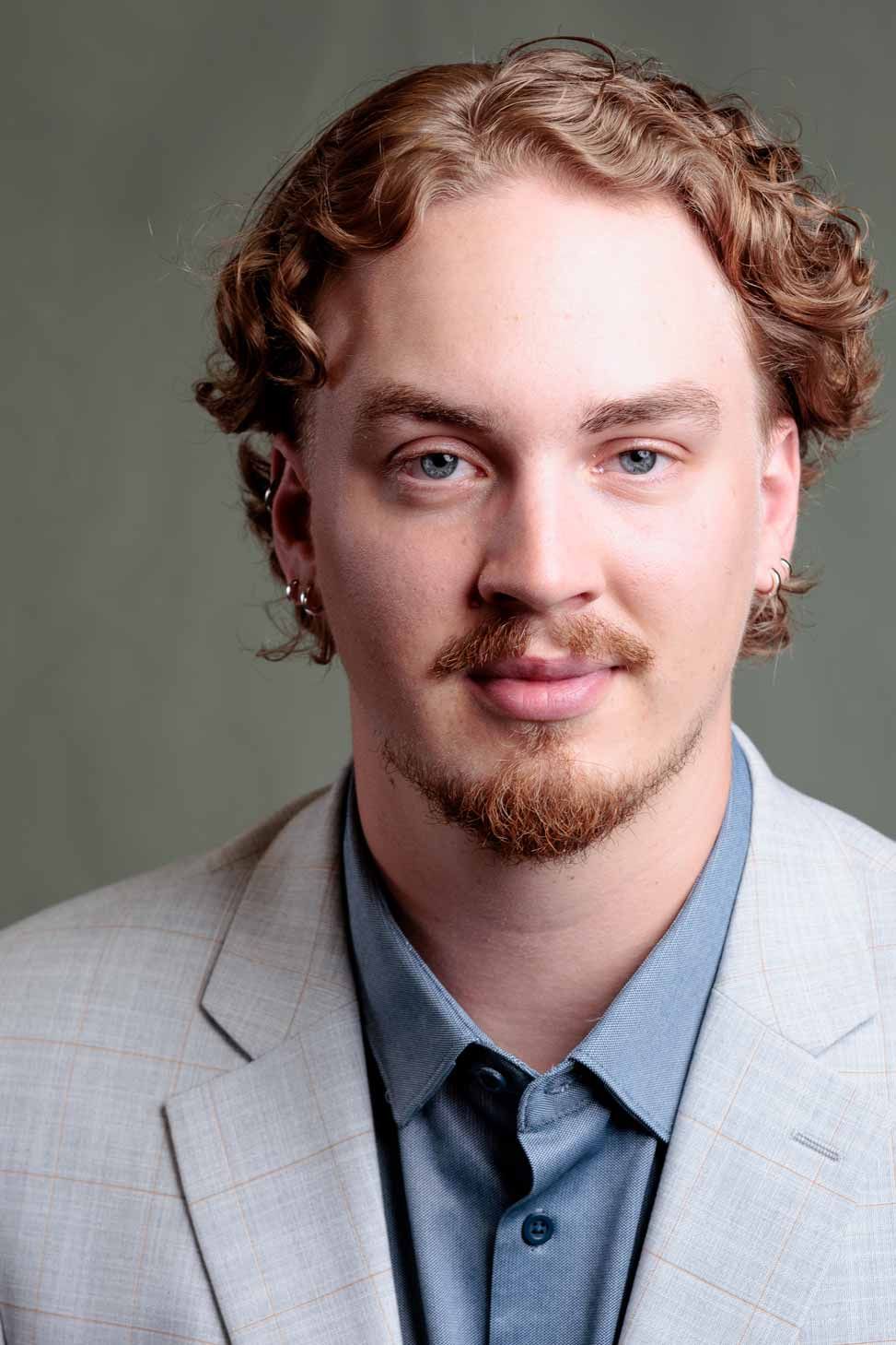 Corporate headshot of man with curly auburn hair wearing gray suit and blue shirt