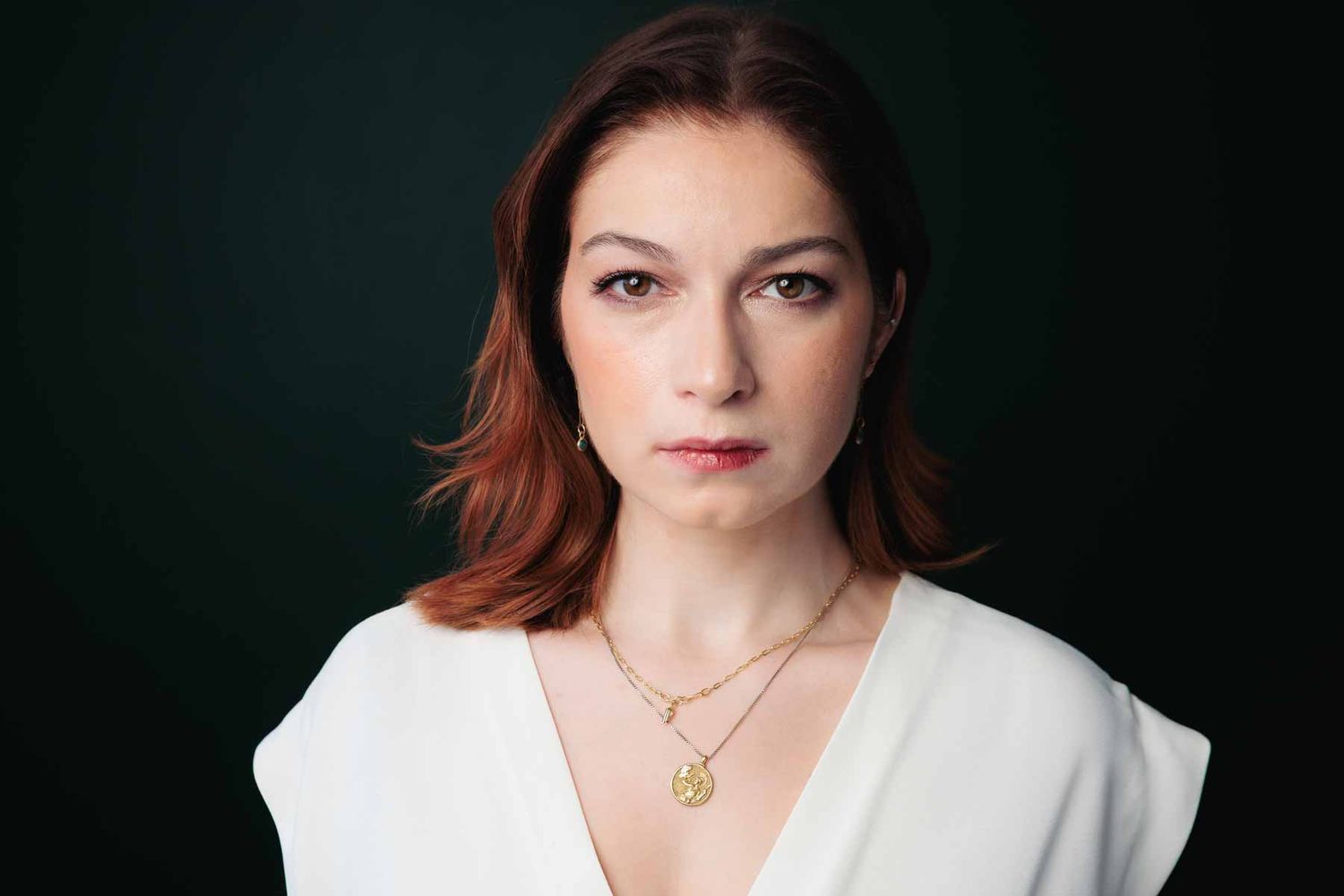 Professional headshot of woman with auburn hair wearing white blouse and gold jewelry against dark background