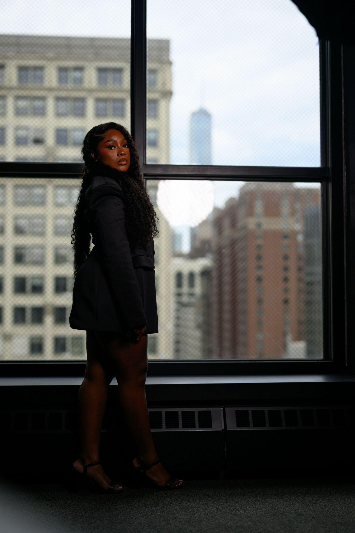 A woman in a black blazer and shorts stands by large windows with a city skyline backdrop.