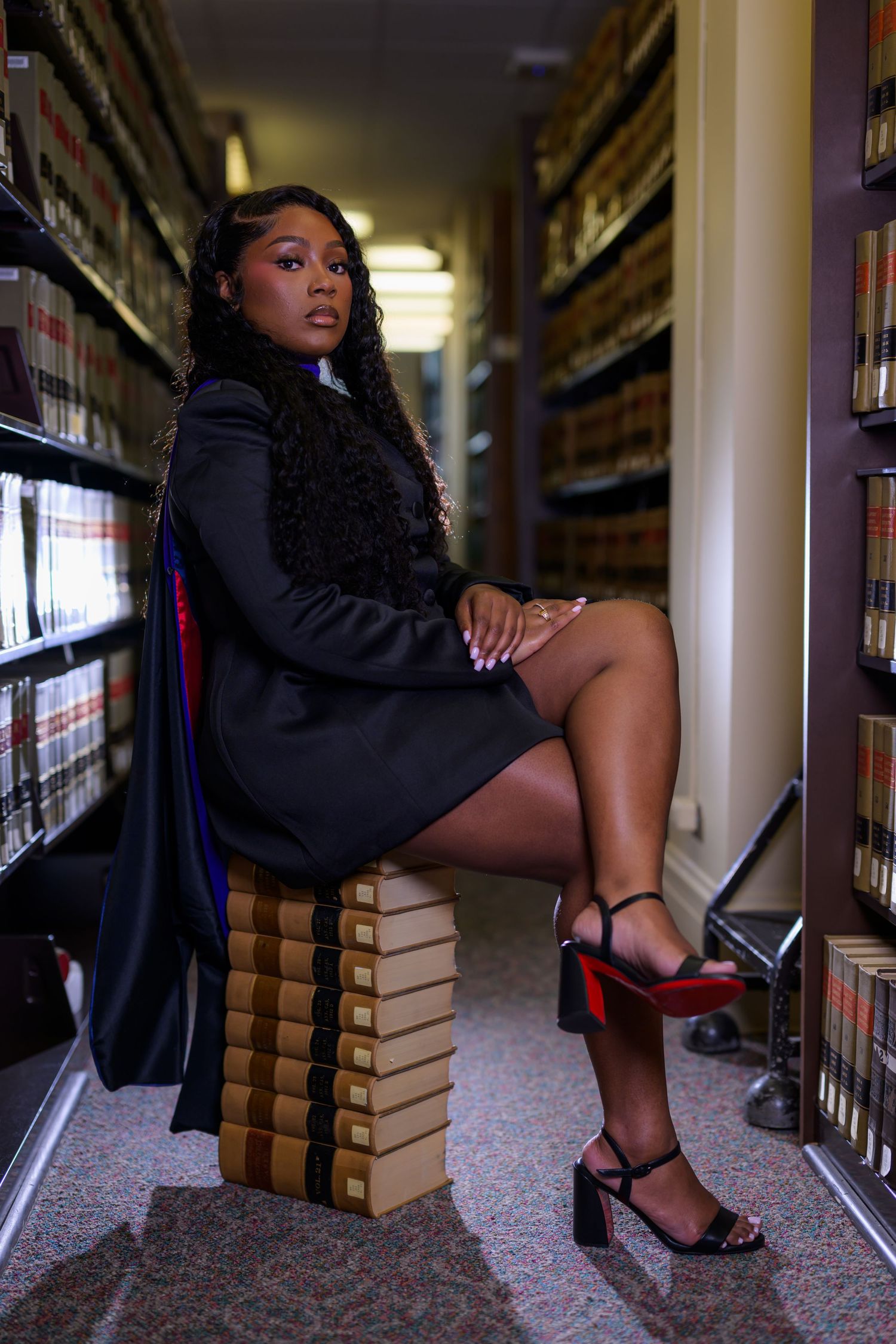 Young woman in black blazer and heels sits on stacked books in a law library aisle.