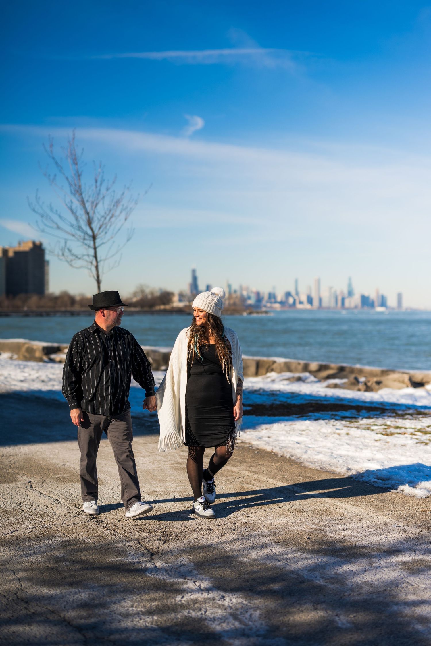 Couple holding hands walking along snowy lakeside path with city skyline in background on sunny winter day.