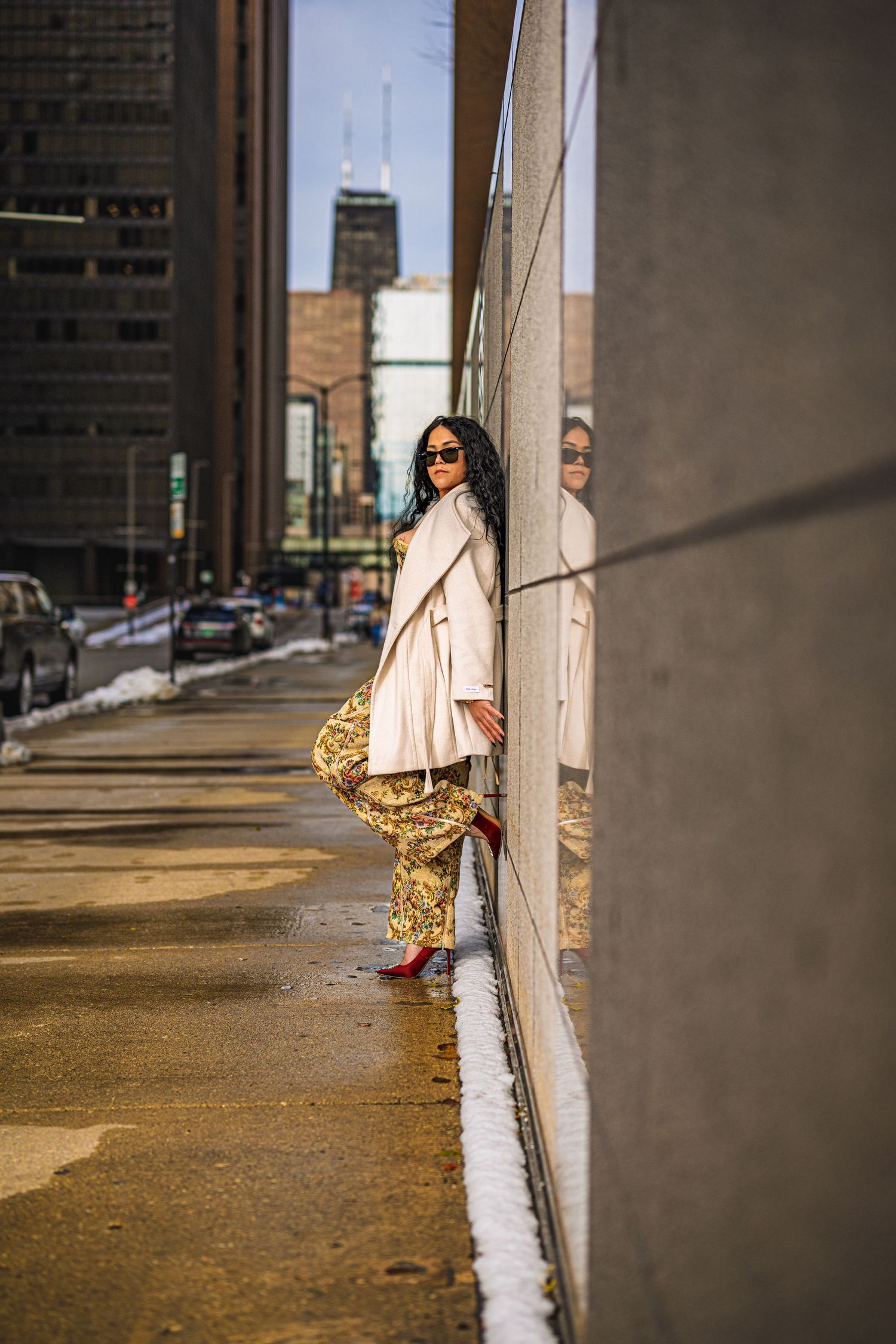 A stylish woman in floral pants, cream coat, and red heels leans against a mirrored wall on a urban city sidewalk.