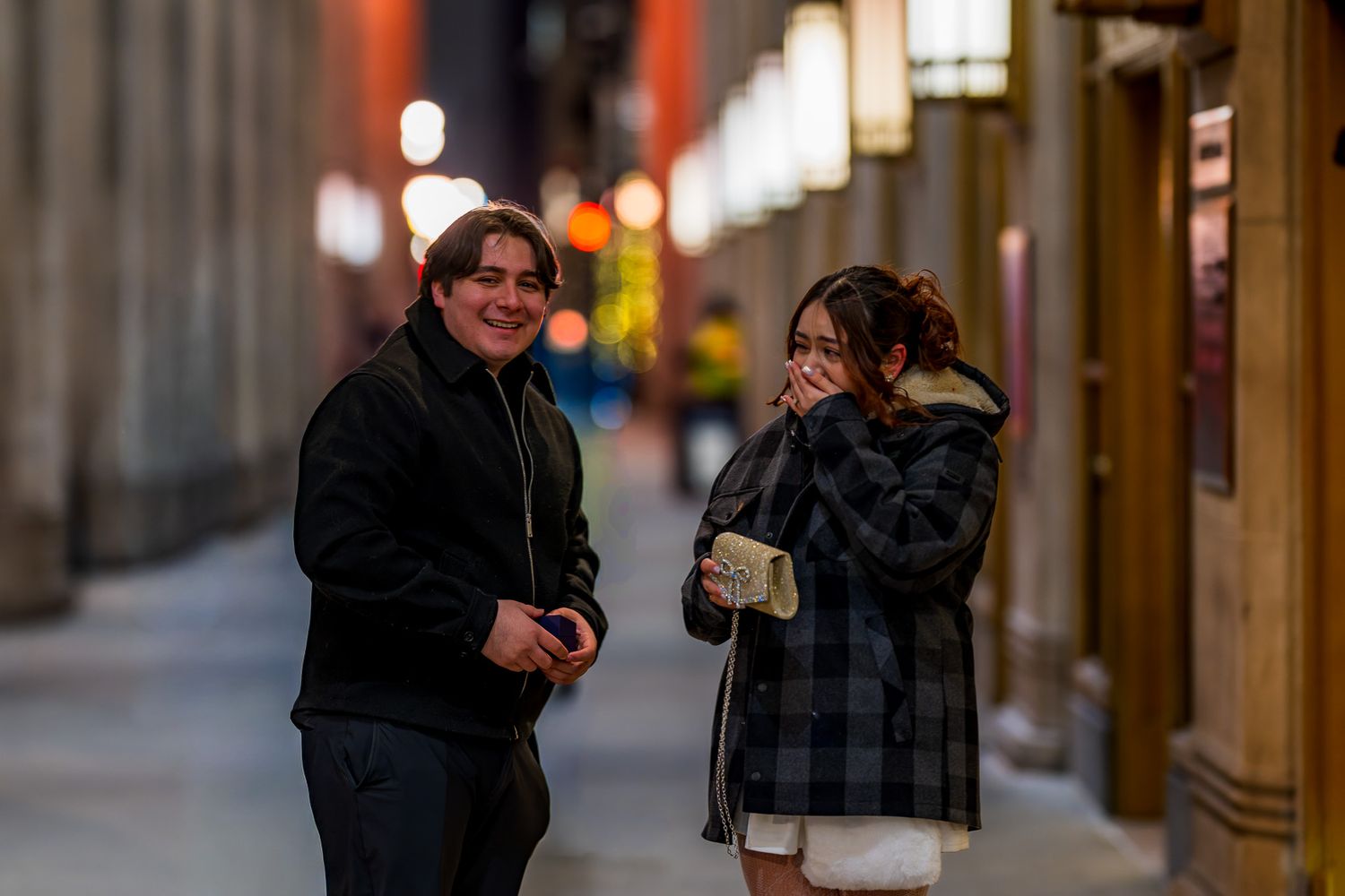 Two young people laughing together on a city street at night, wearing winter coats in an urban alley with bokeh lights.