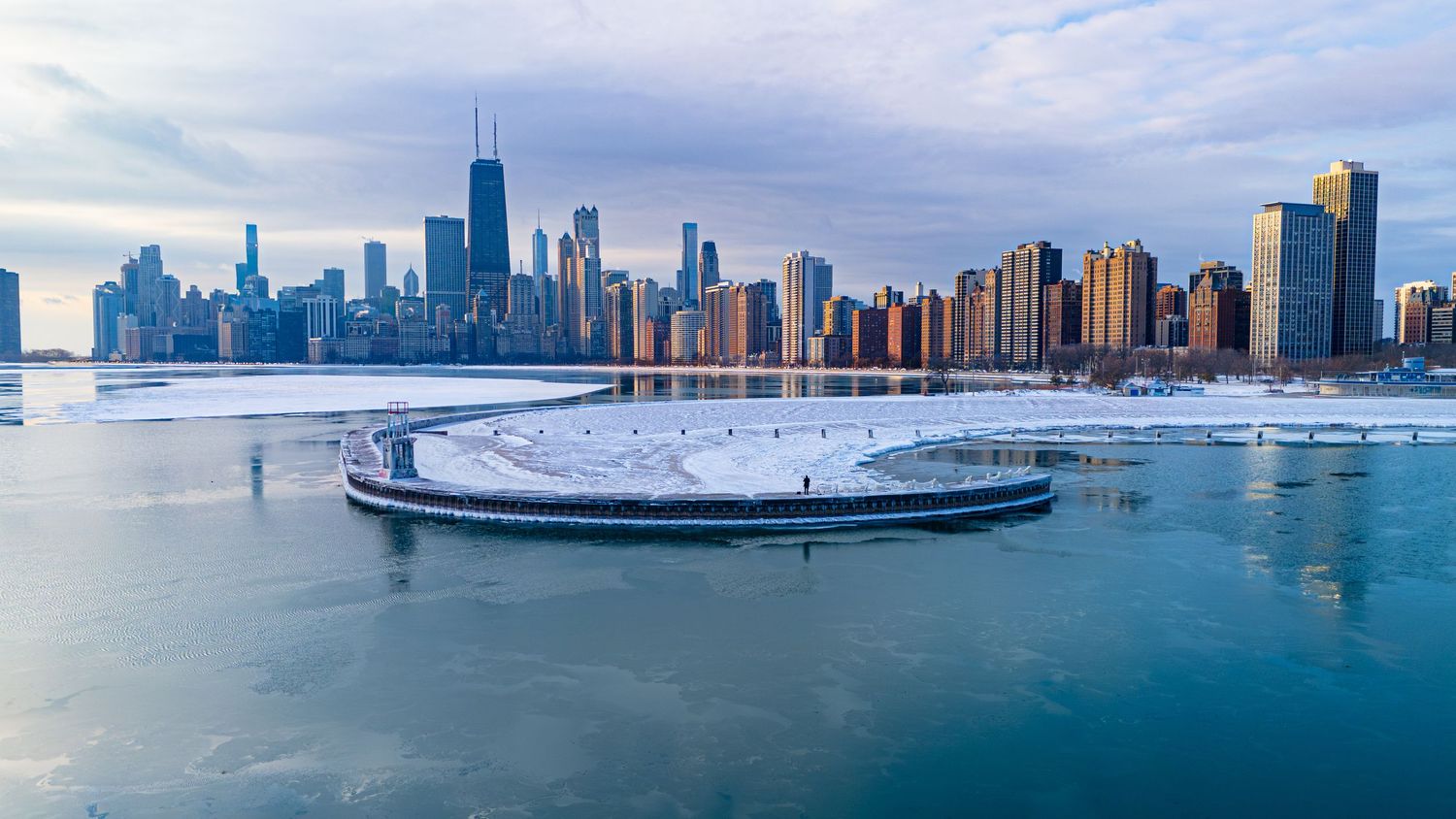 Aerial view of Chicago skyline in winter with frozen Lake Michigan and a snow-covered circular pier in the foreground.