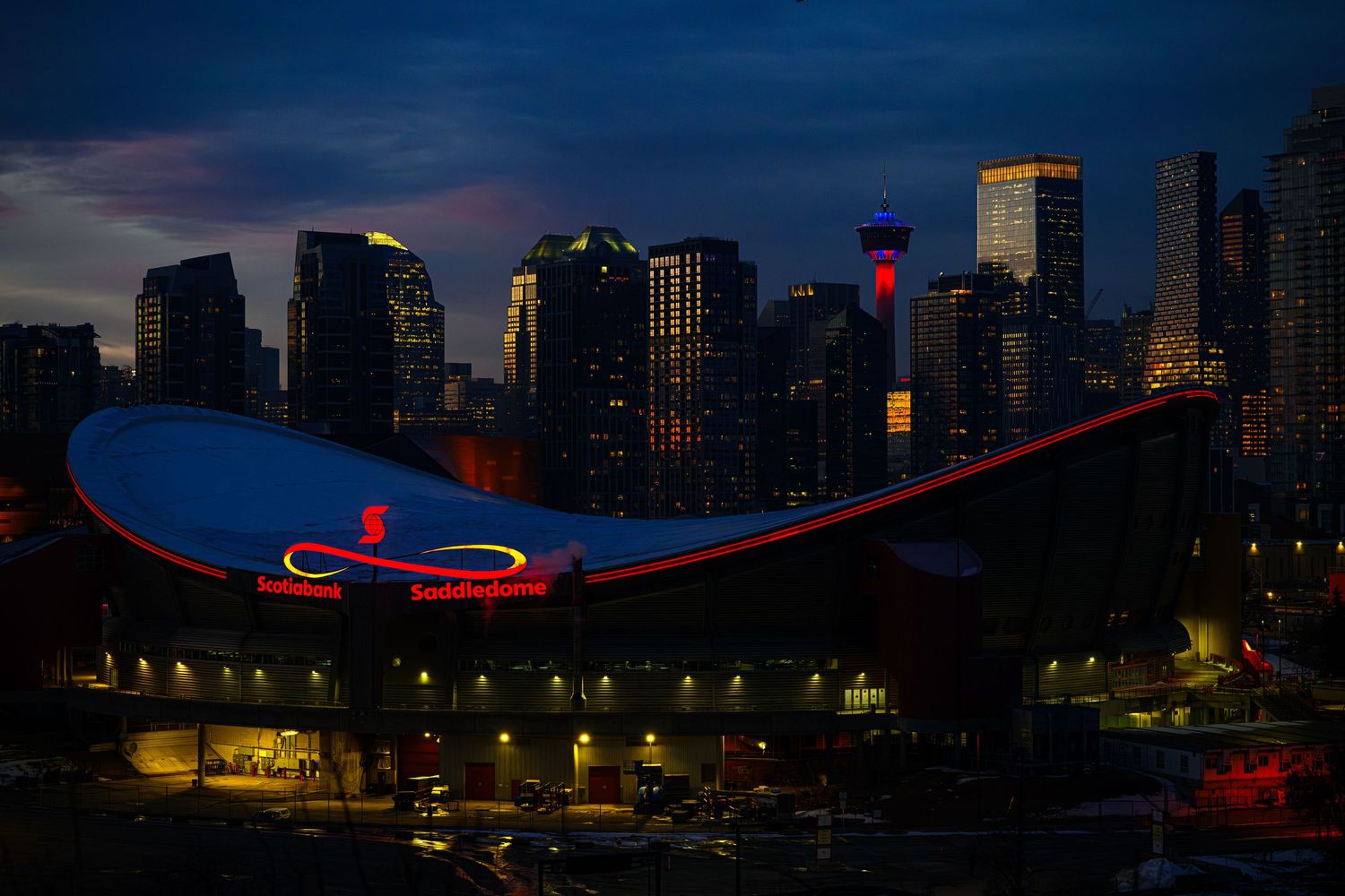 Calgary skyline at dusk with the Scotiabank Saddledome arena illuminated in red and blue lights.