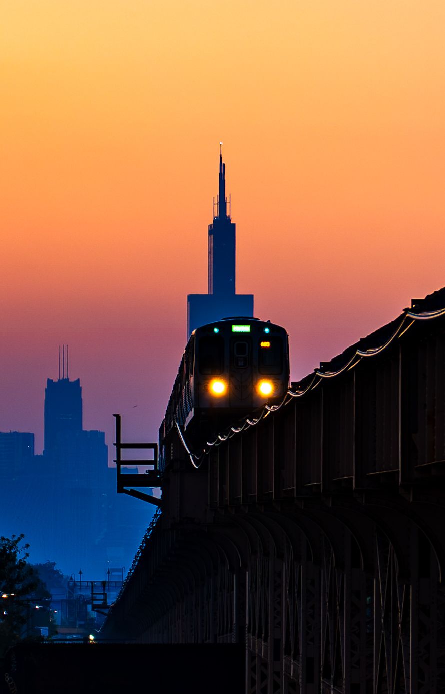 A silhouetted elevated train approaches at sunset with Chicago skyline skyscrapers glowing against an orange and blue dusk sky.