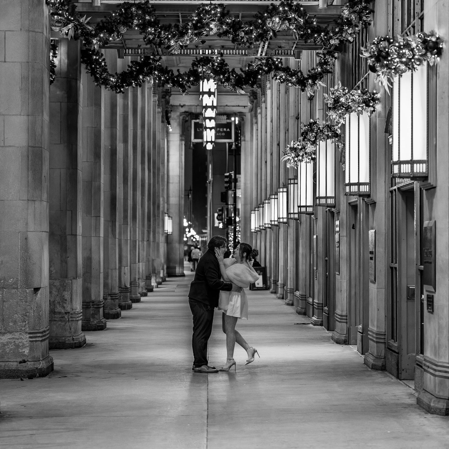 Black and white photo of a couple embracing in an elegant columned arcade decorated with holiday garland.