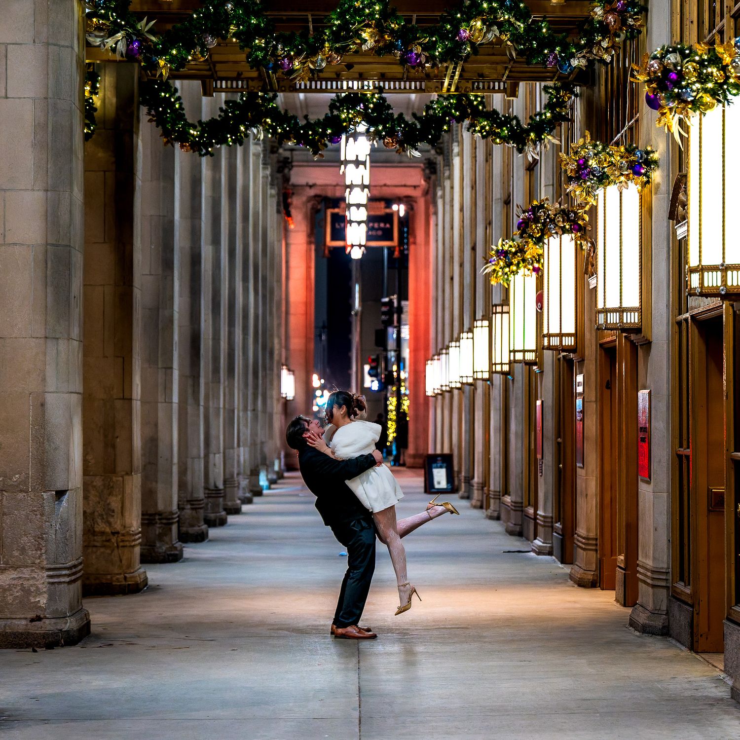 Couple sharing a romantic dip kiss in an elegant columned hallway adorned with festive holiday greenery and warm lights.