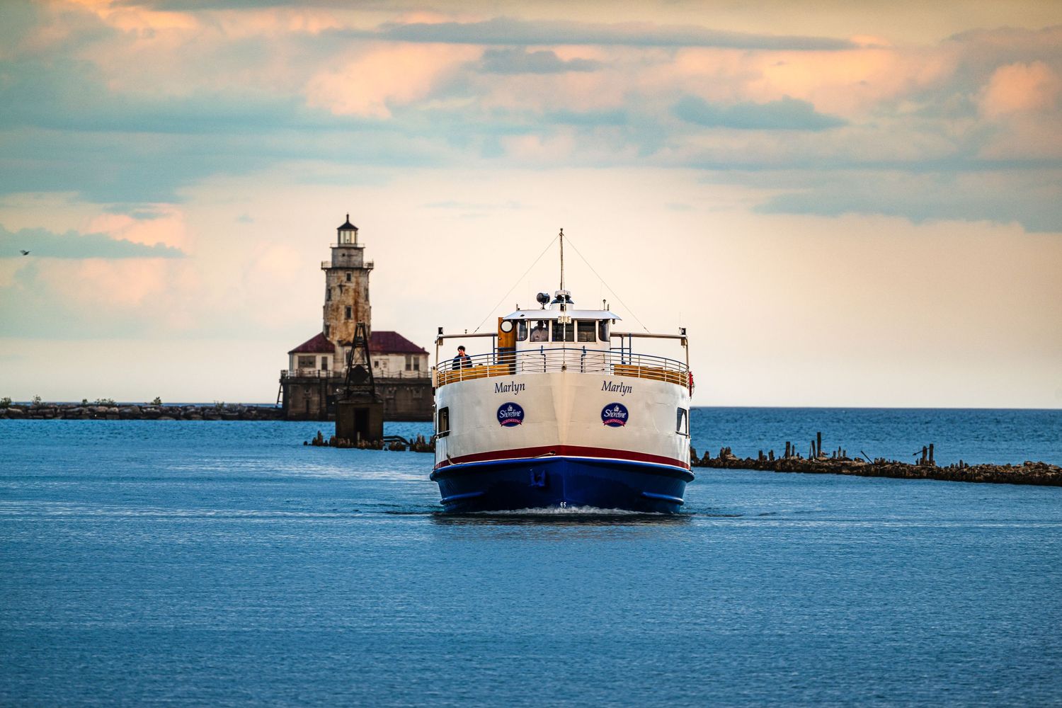 White passenger ferry boat cruising on blue waters near a historic lighthouse under a dramatic cloudy sunset sky.