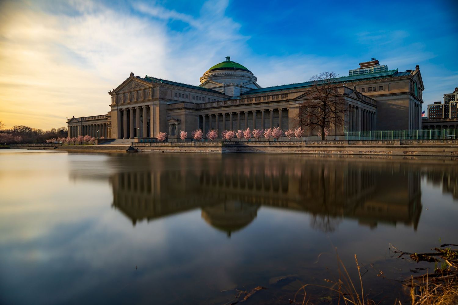Museum of Science and Industry in Chicago reflects in calm water at sunset with American flags and dramatic sky.