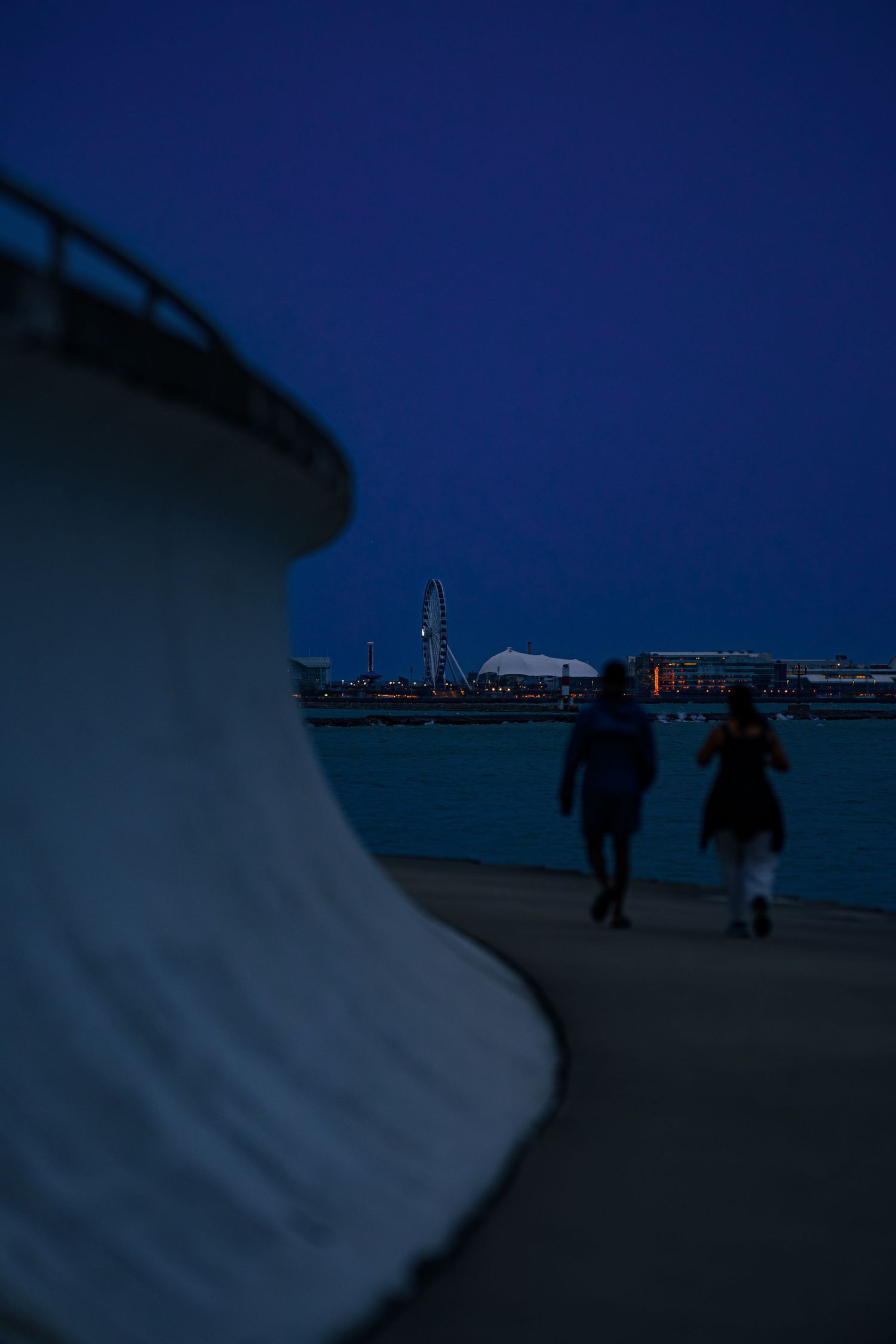 Two silhouetted figures walk along a waterfront promenade at dusk with a illuminated cityscape in the background.