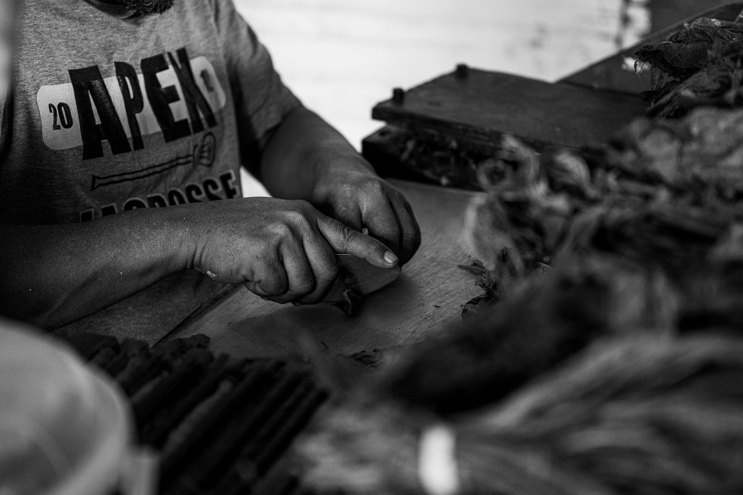 Man in APEX t-shirt hand-rolling cigars on a workbench in a black and white photograph.
