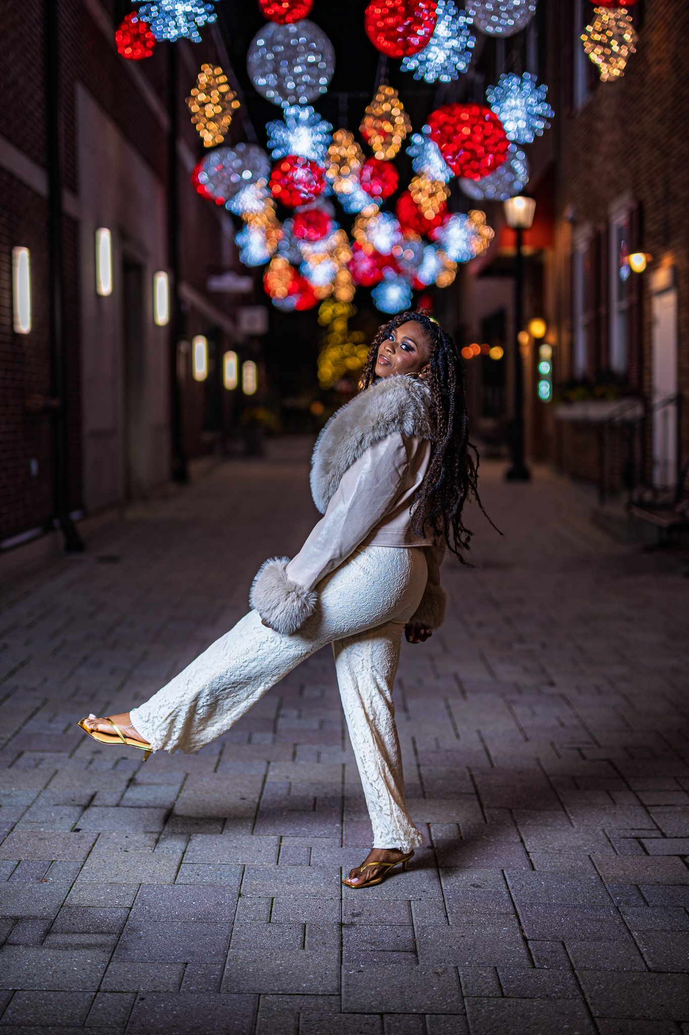 Woman in white outfit with fur trim poses on cobblestone street under colorful holiday light decorations at night.