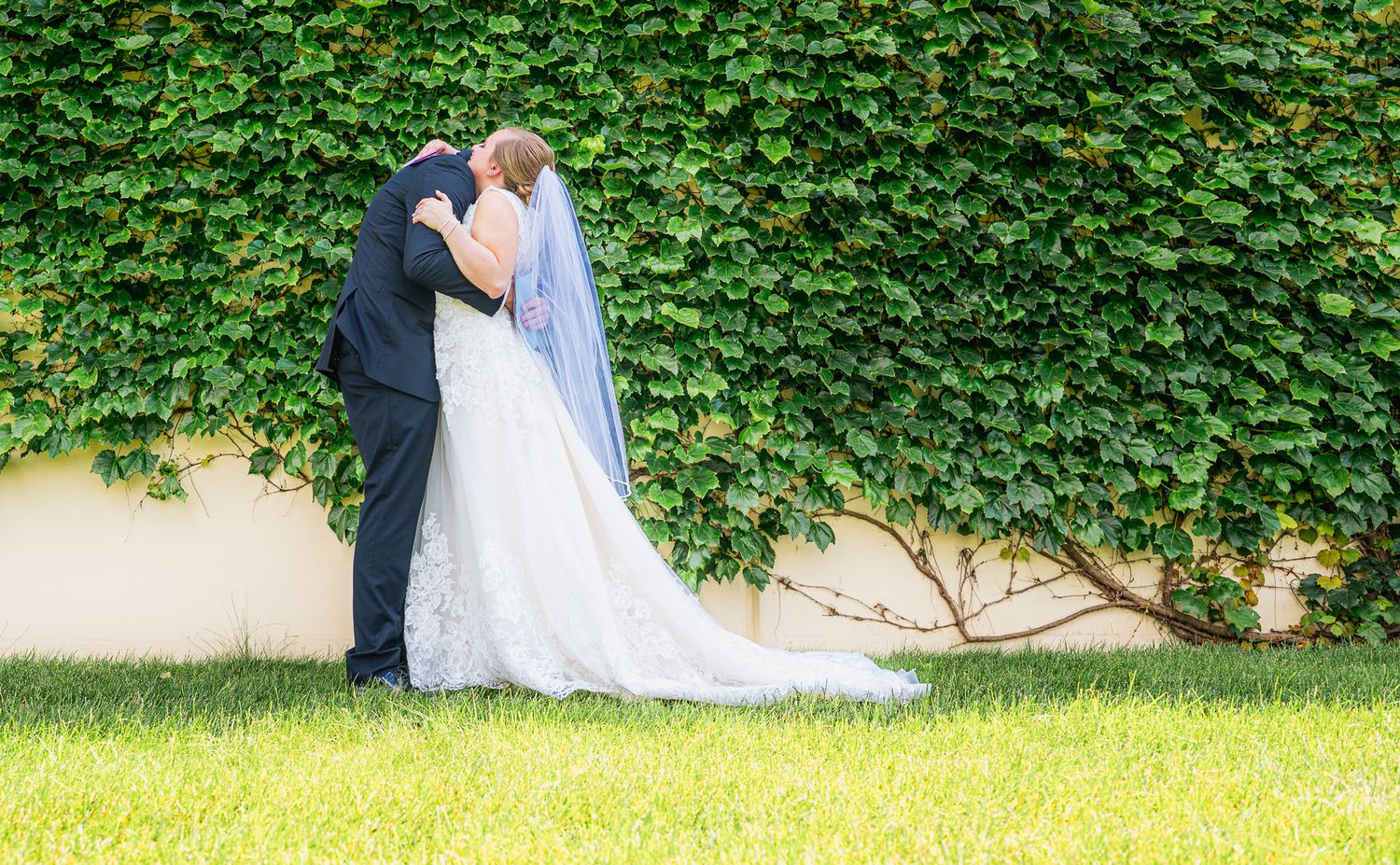Bride and groom share a romantic kiss in front of a lush ivy-covered wall on their wedding day.