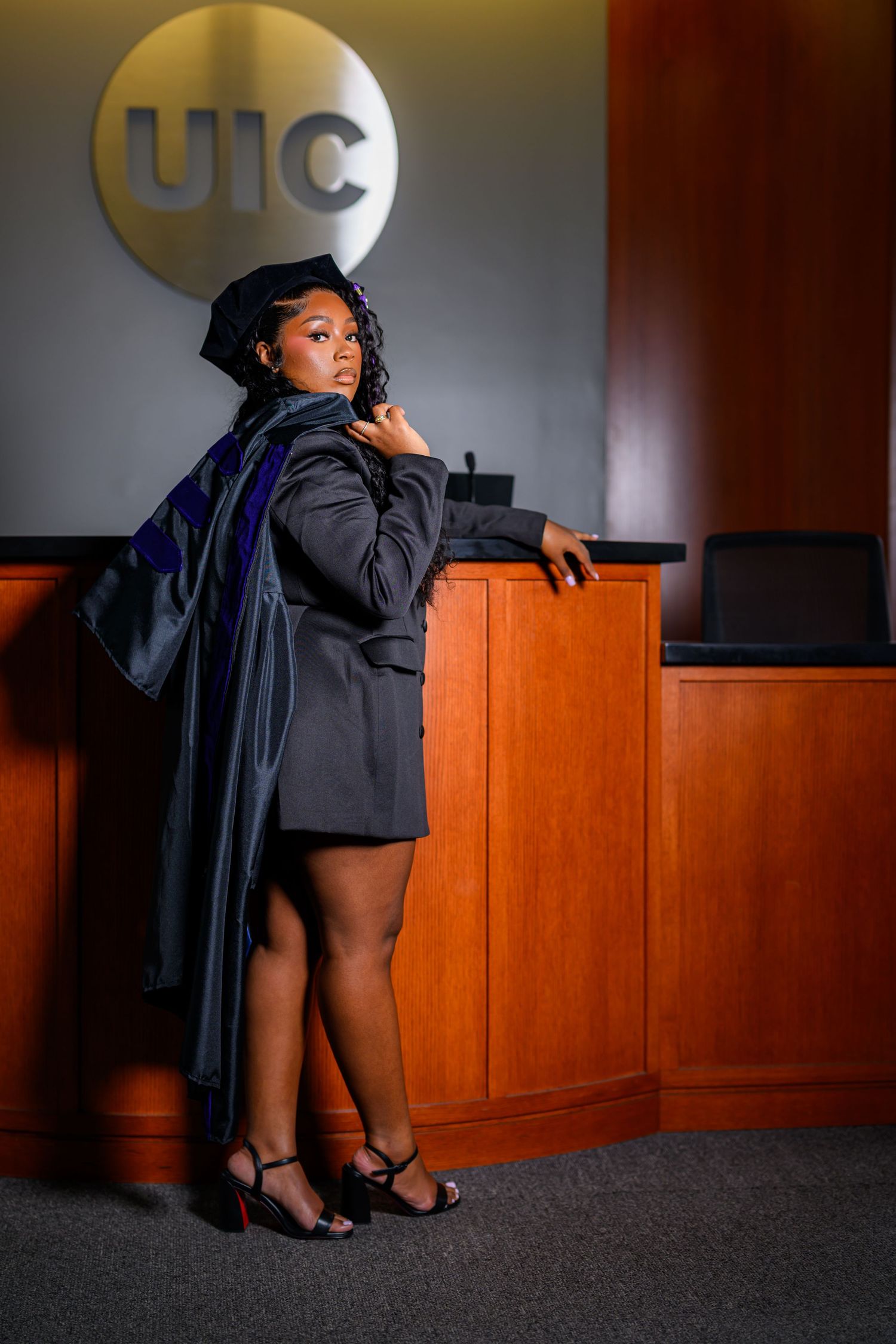 A proud graduate poses in cap and gown at UIC, wearing heels and draping her academic regalia over her shoulder.