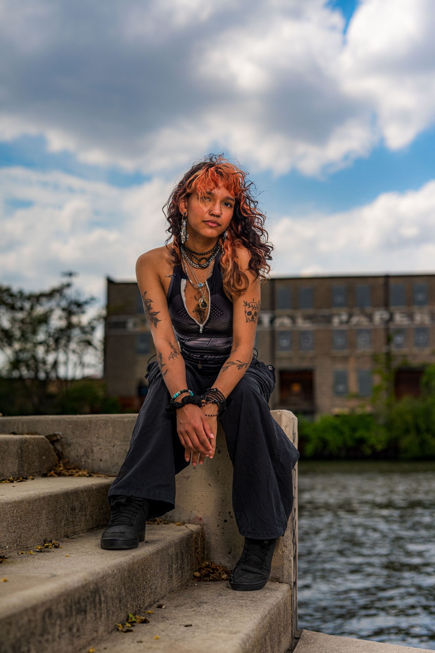 Young woman with curly red hair and tattoos sits on concrete steps by a river, wearing dark casual outfit.