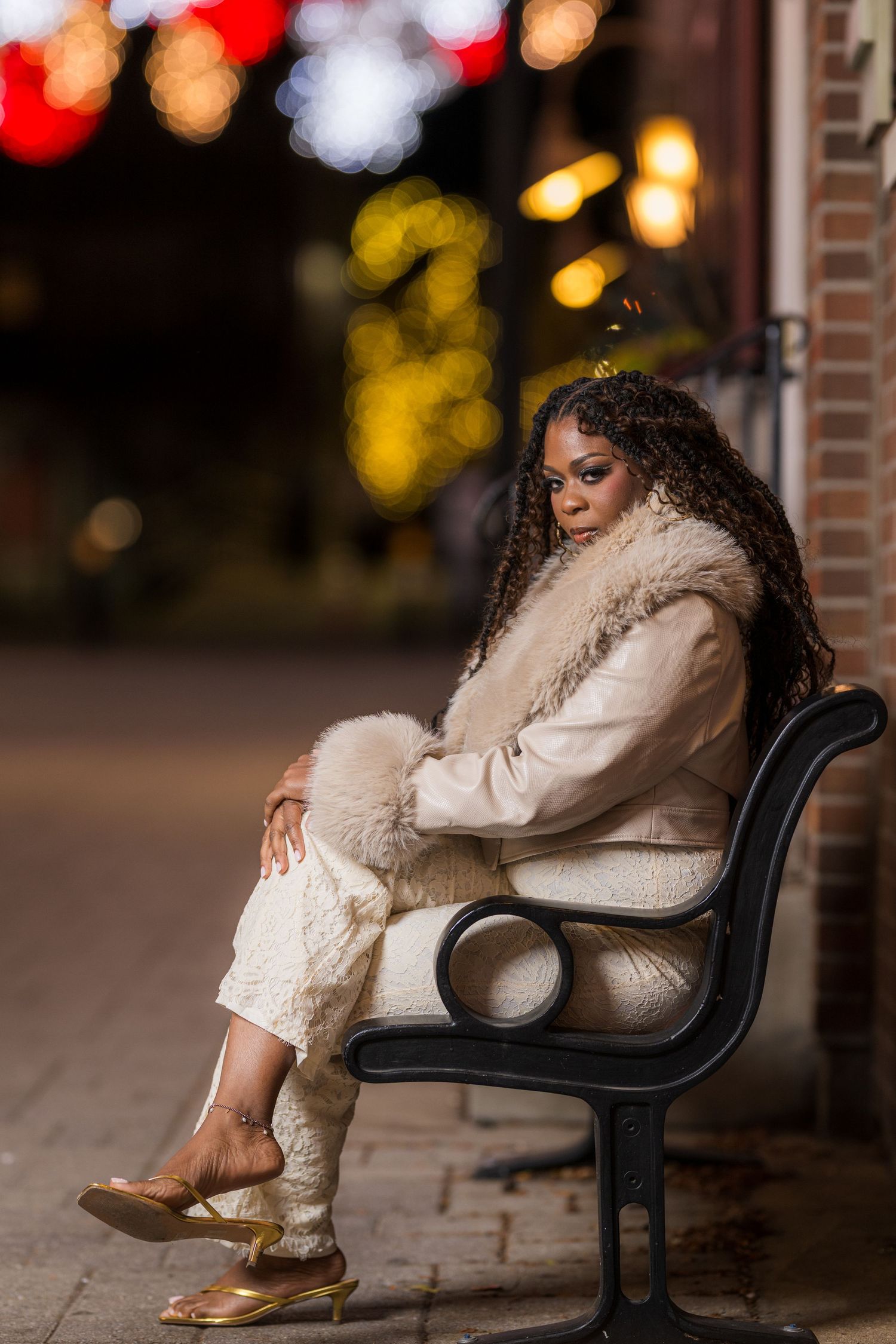 A Black woman in a cream fur coat and gold heels sits elegantly on a bench at night with bokeh lights behind her.
