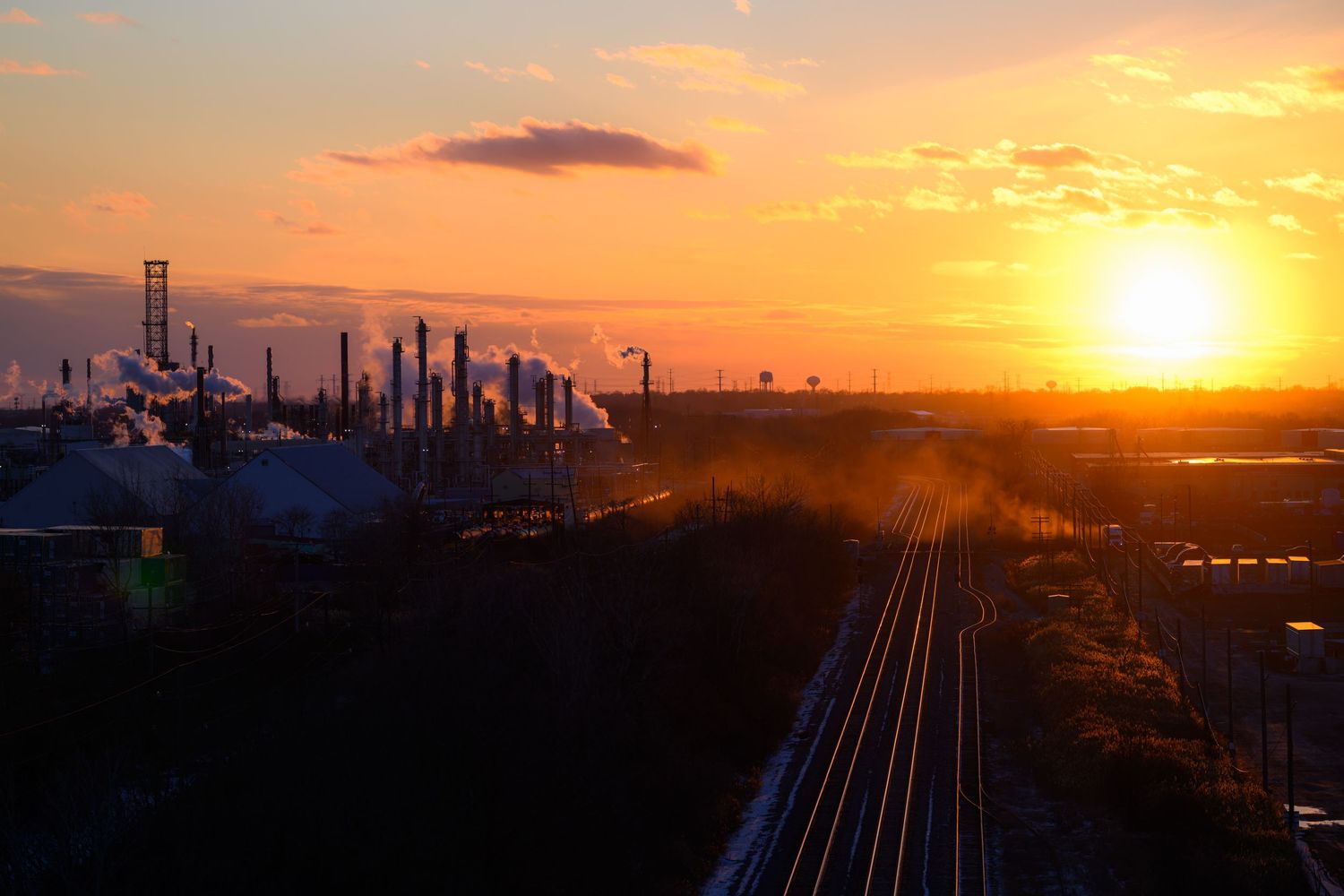 Aerial view of an industrial refinery silhouetted against a vibrant orange and yellow sunset with railroad tracks in foreground.