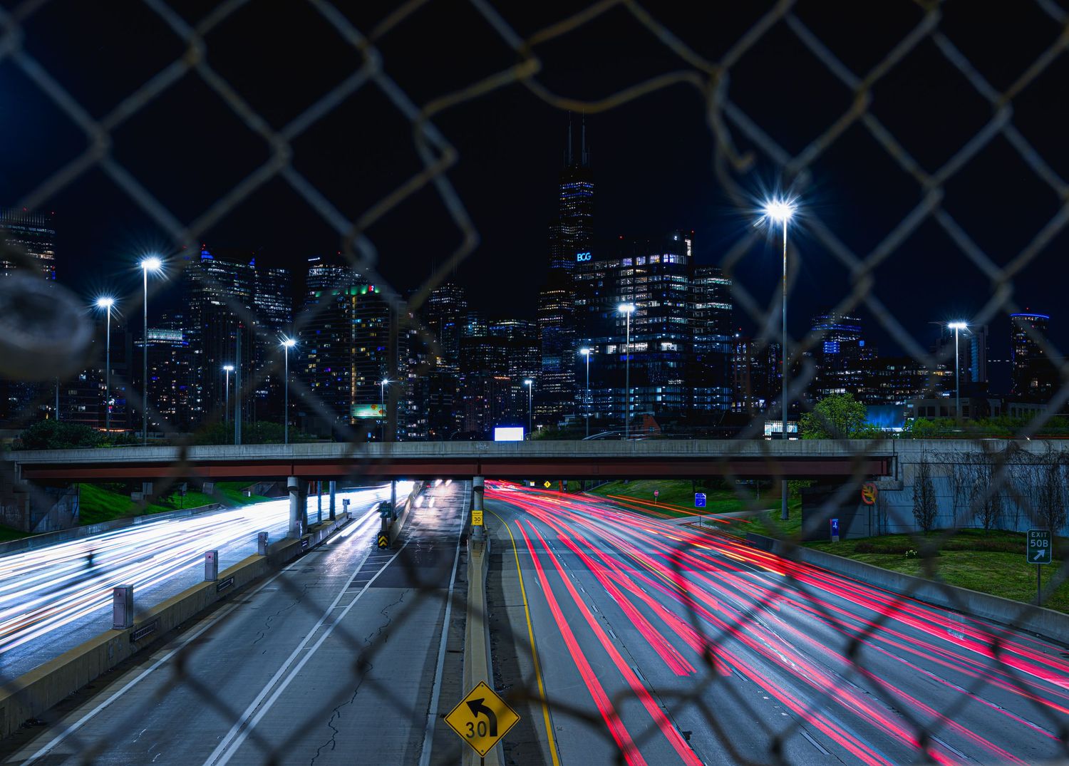 Night city skyline viewed through chain-link fence with light trails on highway below.