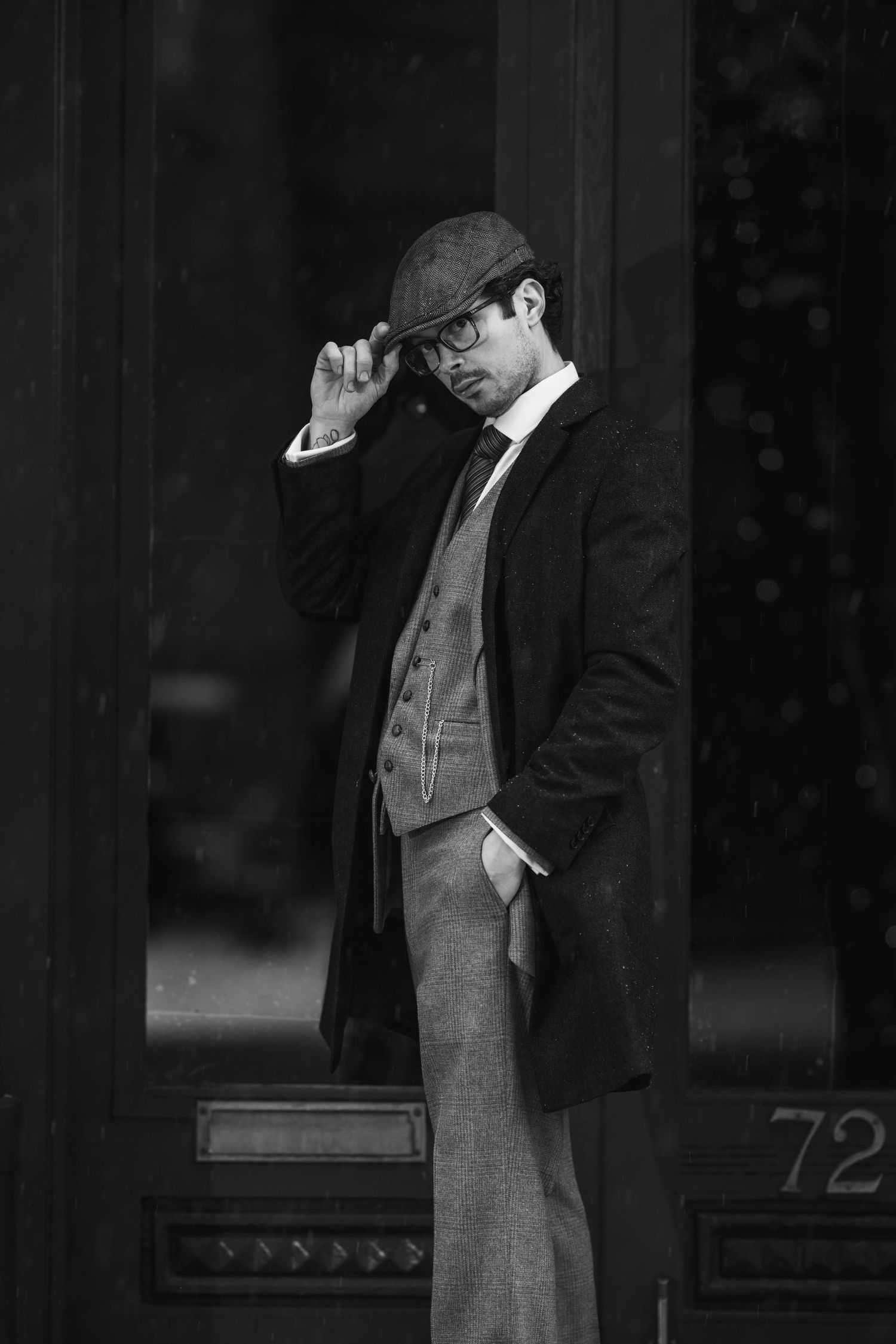 Black and white portrait of a dapper man in vintage suit, overcoat, and flat cap, adjusting his hat outside.