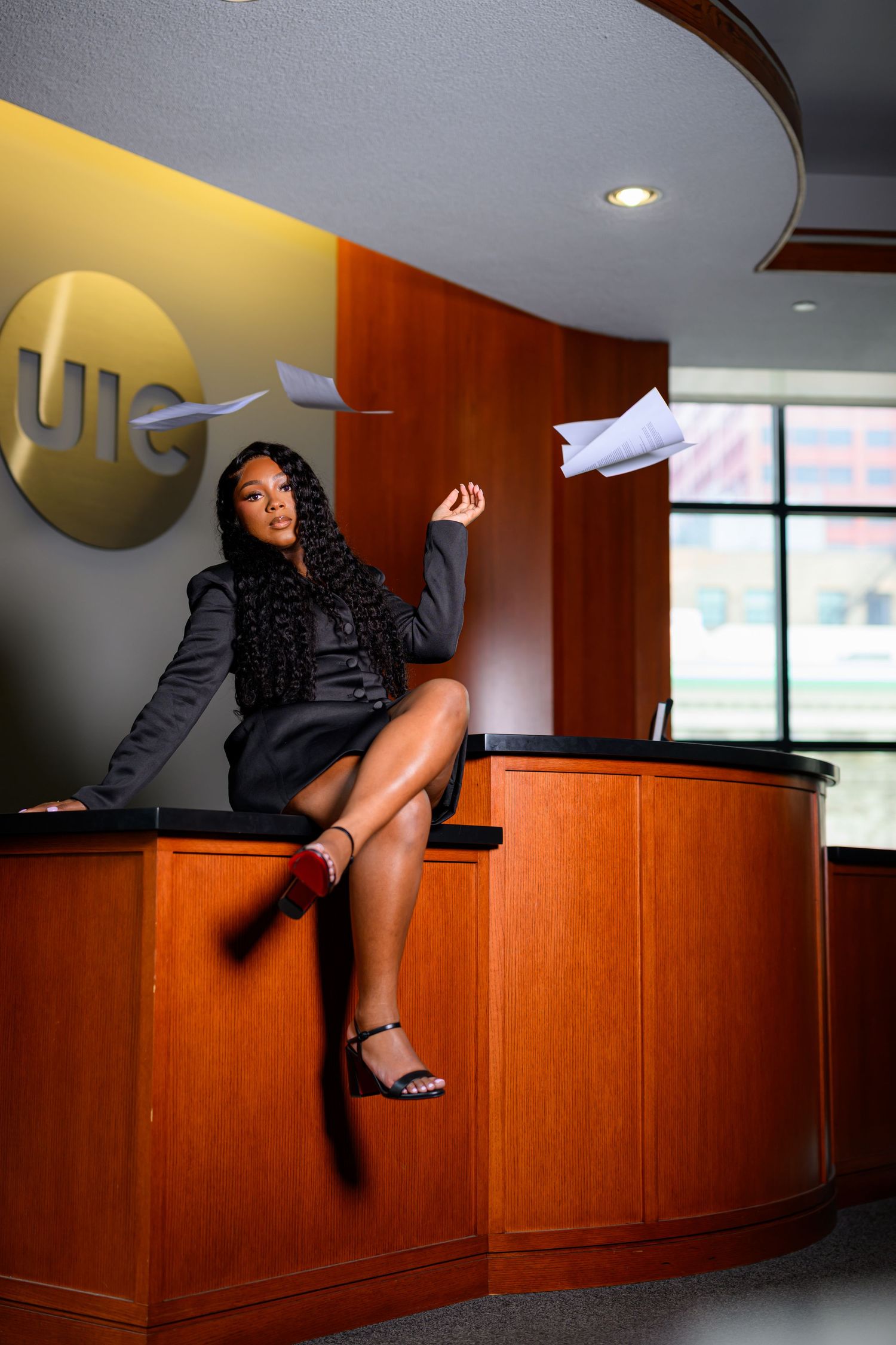 A confident Black woman in a black dress sits on a reception desk, tossing papers in the air in a modern office lobby.