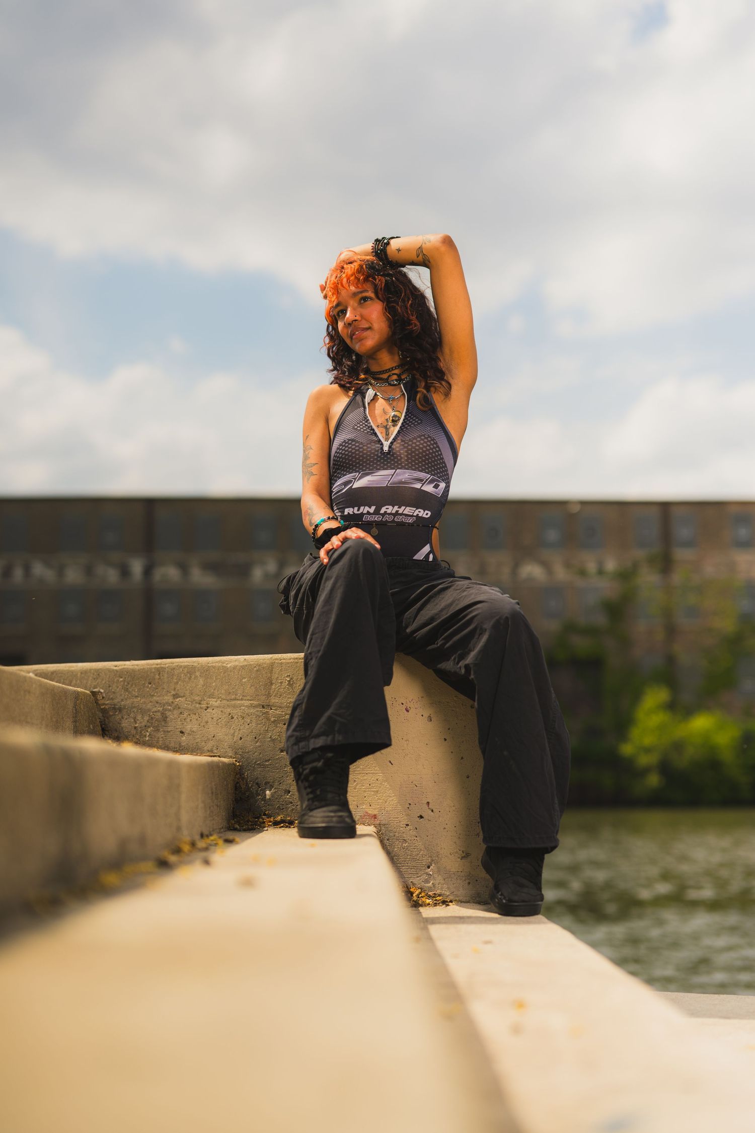 A young woman with red hair sits on concrete steps outdoors, wearing black wide-leg pants and a blue top, posing confidently.