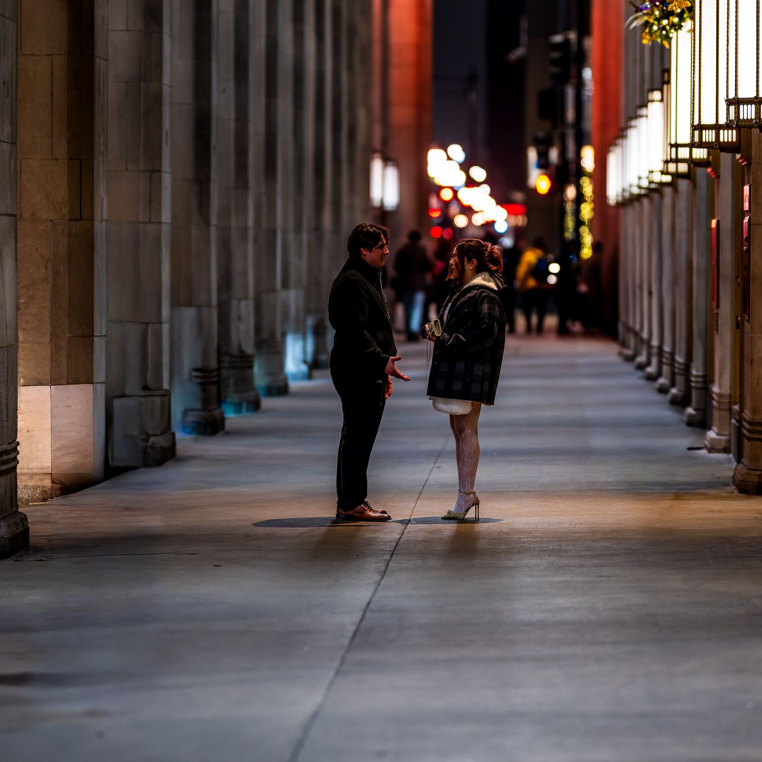 Two people conversing at night under a columned walkway with bokeh city lights in the background.