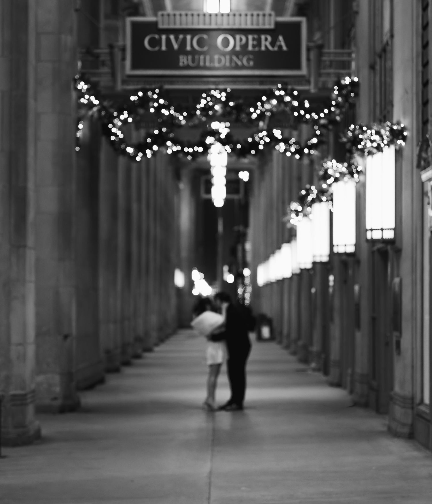 Black and white photo of a couple embracing in the Civic Opera Building arcade, adorned with twinkling holiday lights.