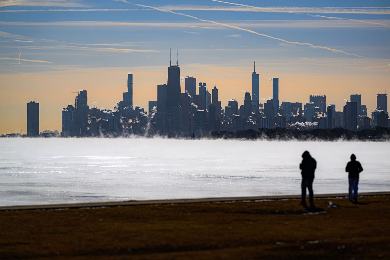 Two silhouetted figures stand on a beach overlooking a misty Chicago skyline at sunset with fog rolling off Lake Michigan.