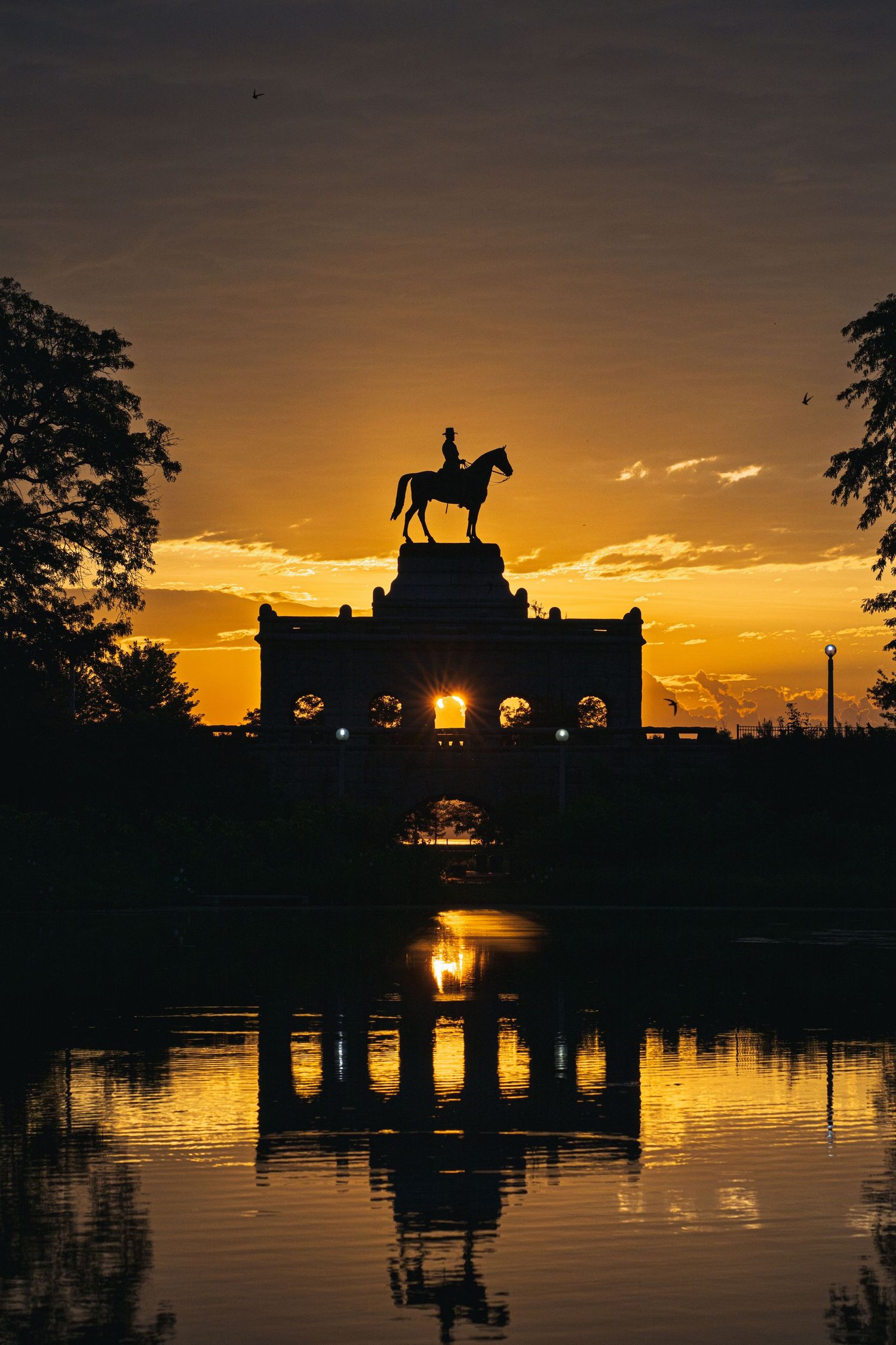 Silhouette of equestrian statue atop archway reflected in pond at golden sunset in a park.