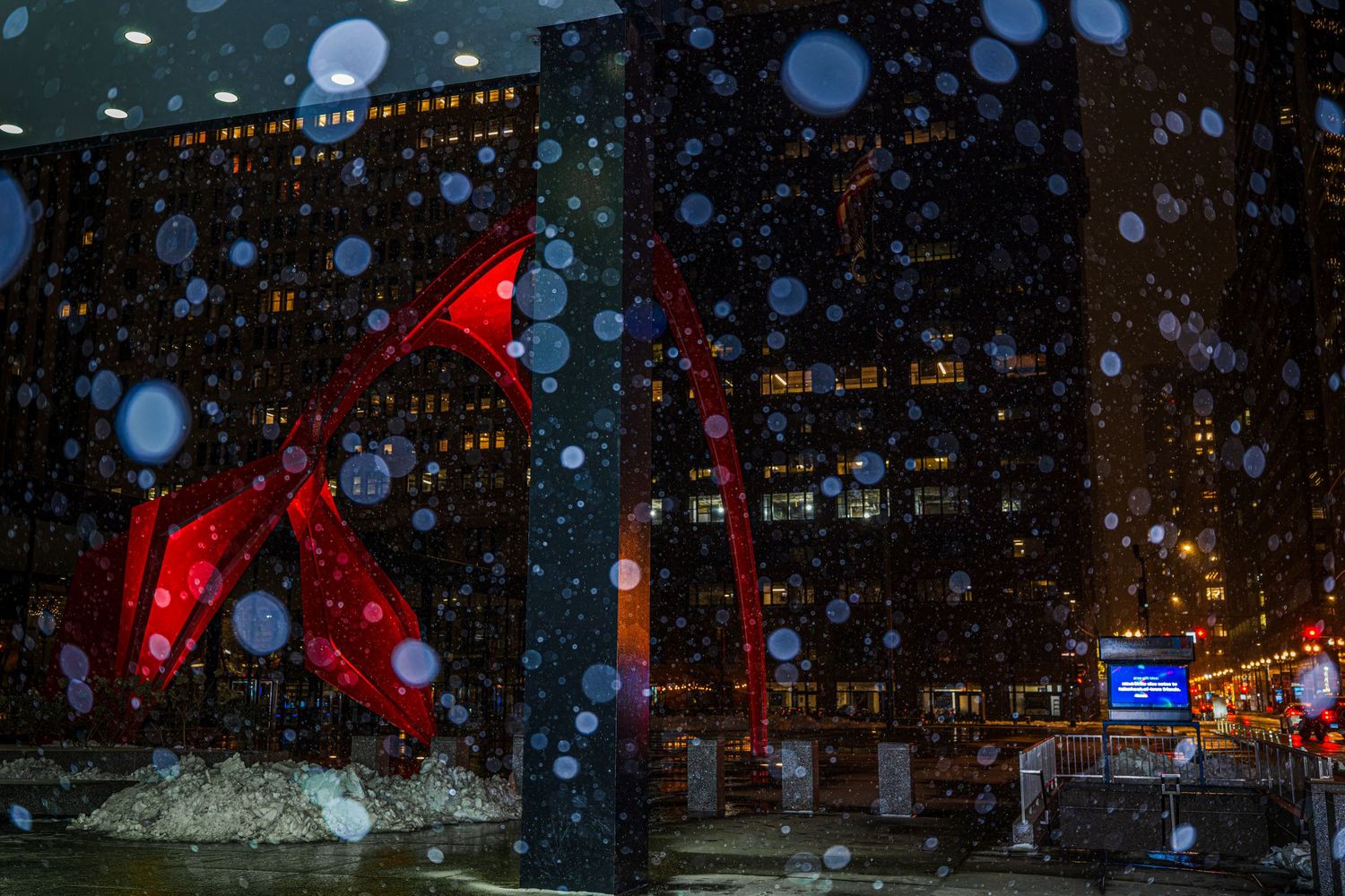 Red sculpture glows in snowy night cityscape with bokeh lights and dark urban buildings in background.