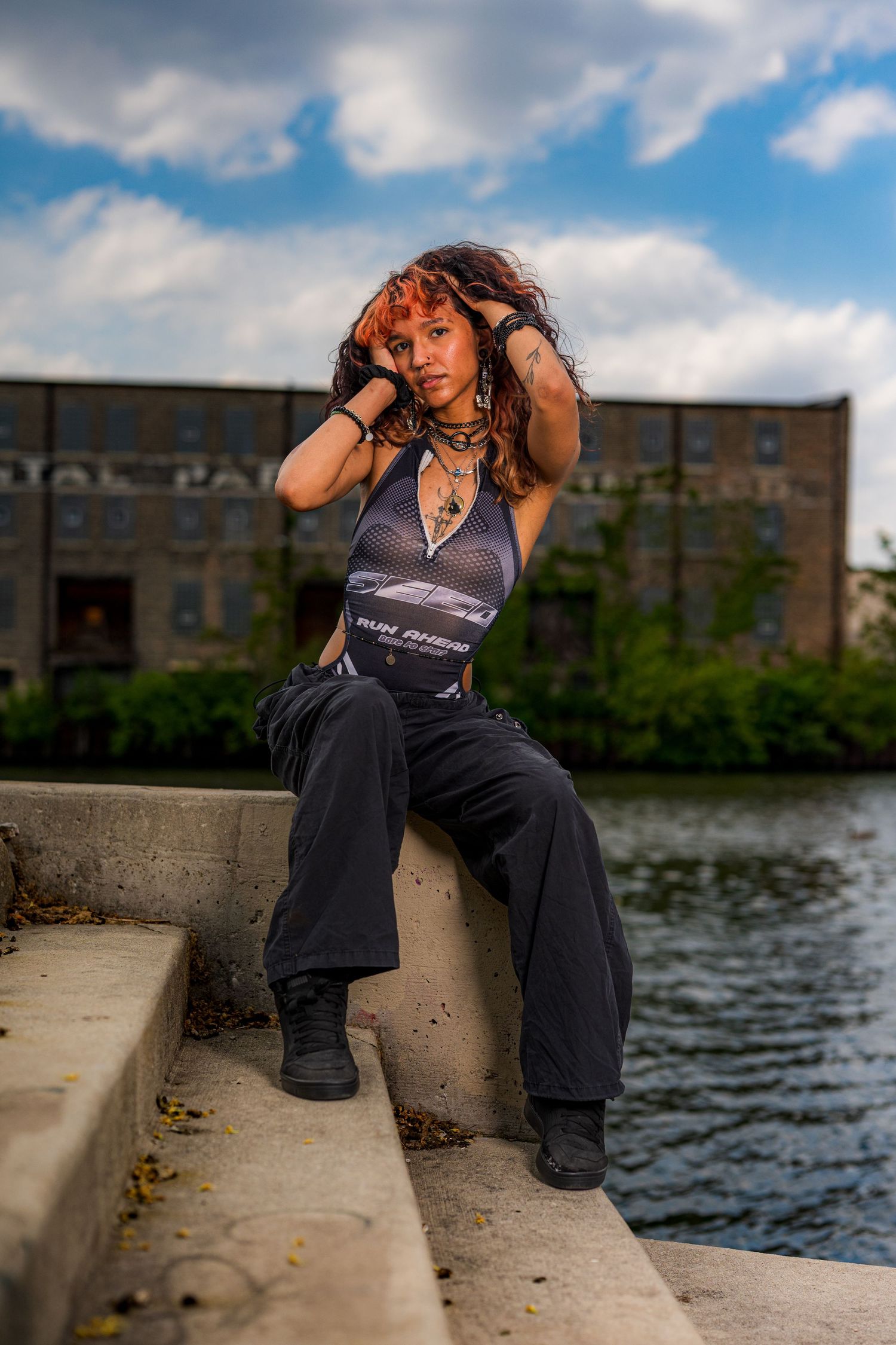 Young woman with red hair poses on concrete steps by a river, wearing a mesh bodysuit and black wide-leg pants.