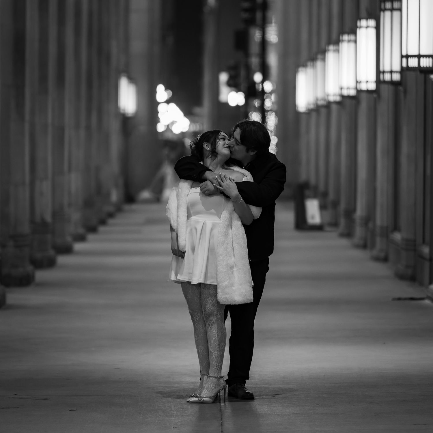 Black and white photo of a couple sharing a romantic kiss in an illuminated colonnade at night.