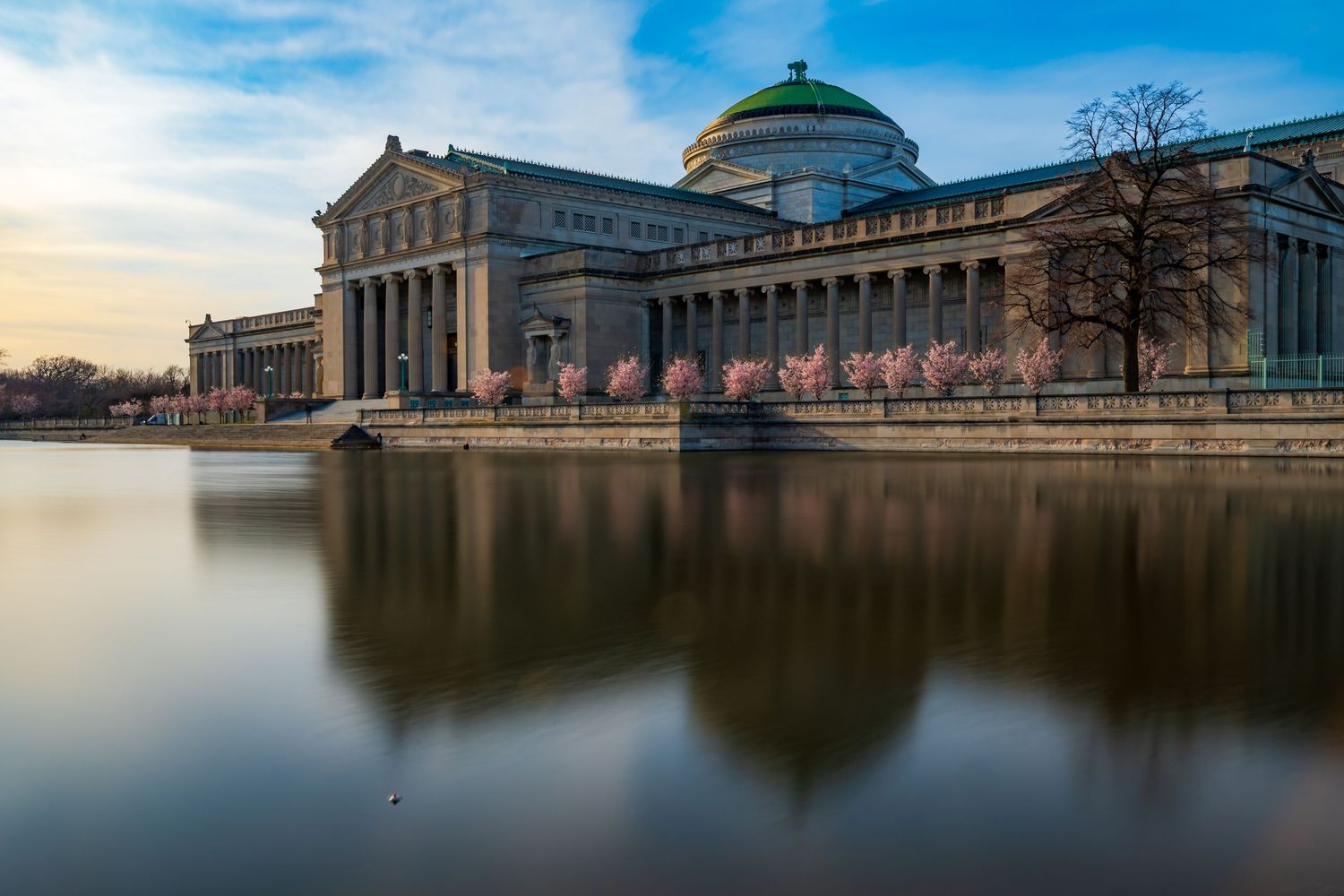 Grand neoclassical museum with green dome reflects in calm water under a blue sky at dusk.