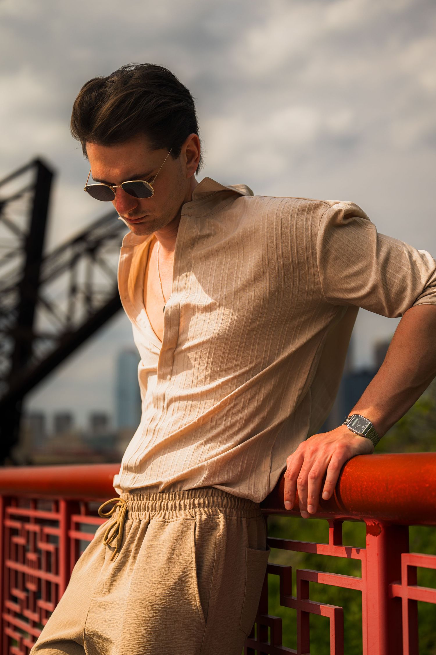 Stylish young man in beige linen outfit and sunglasses leaning on red railing with city skyline backdrop.