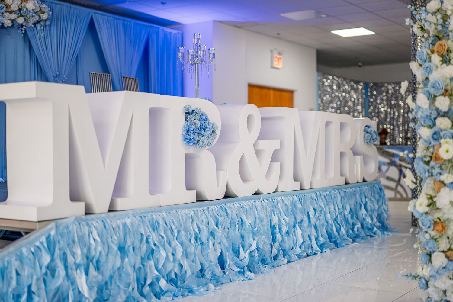 Large white MR & MRS letters on a blue ruffled table at an elegant wedding reception with floral decor.
