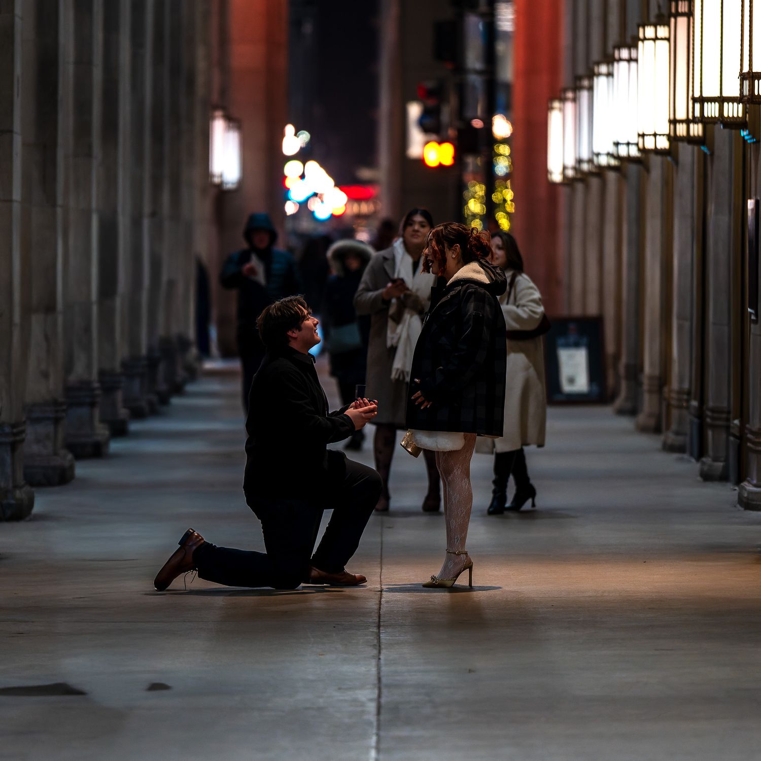 Man proposes on one knee to woman in elegant city arcade at night with onlookers nearby.
