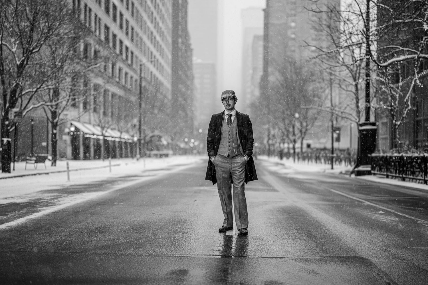 Man in suit and overcoat stands on empty snowy urban street in black and white winter cityscape photo.