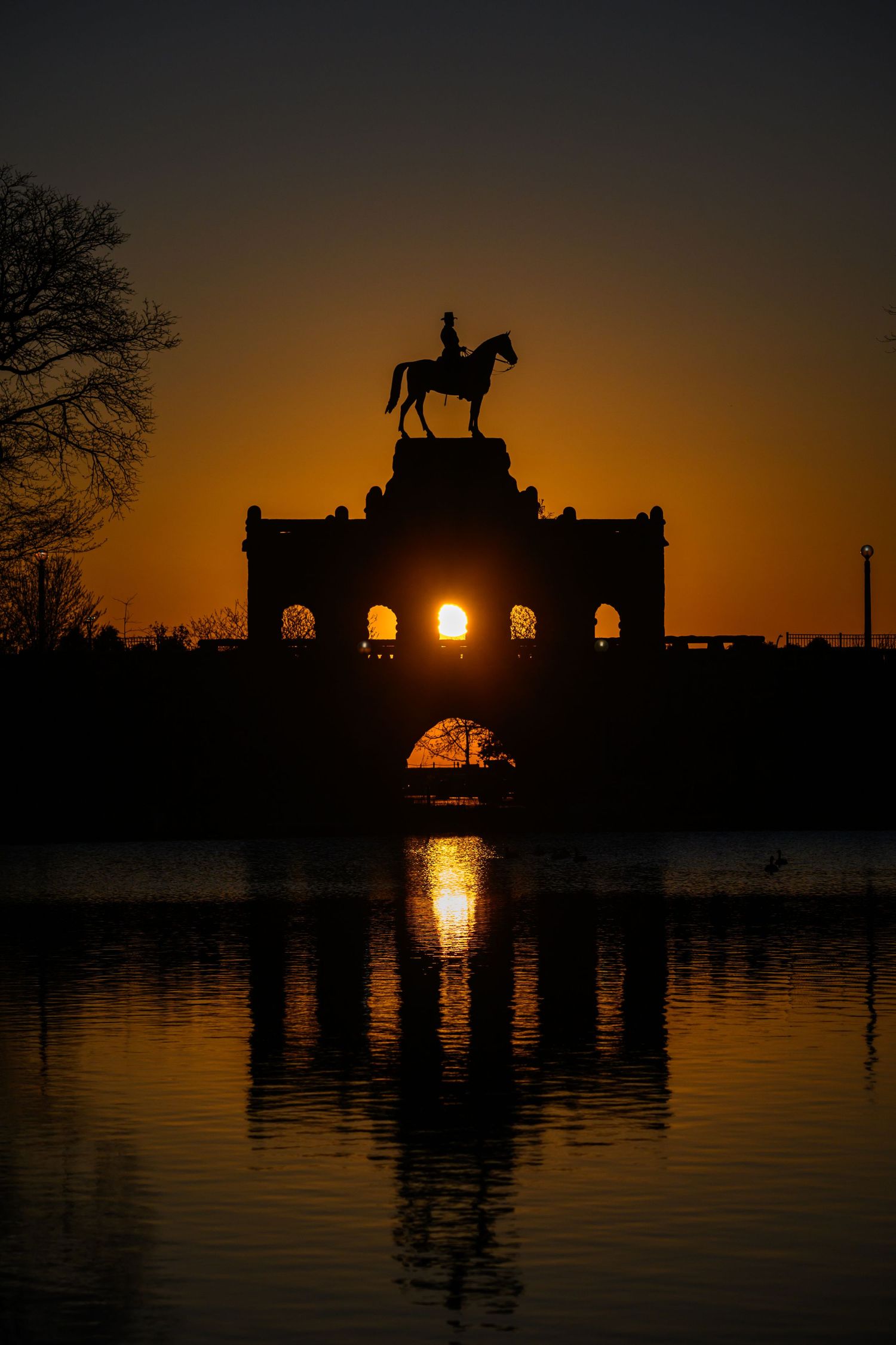 Silhouette of equestrian statue atop stone arch reflecting in water at golden sunset.