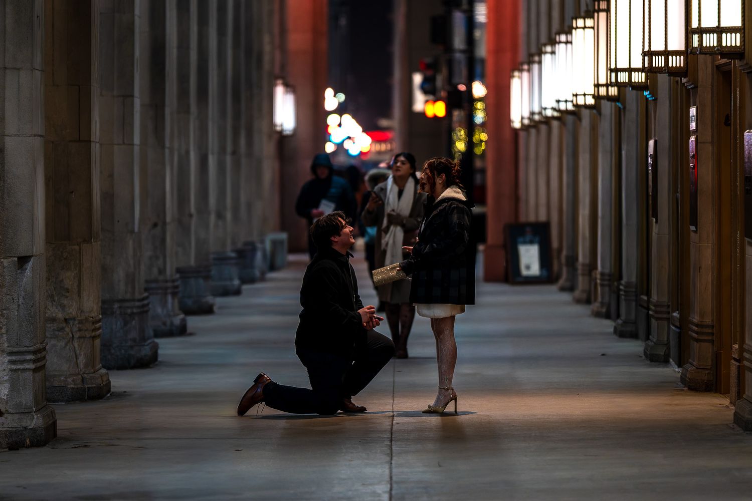 Man proposing on one knee to woman in elegant columned corridor at night, romantic marriage proposal scene.