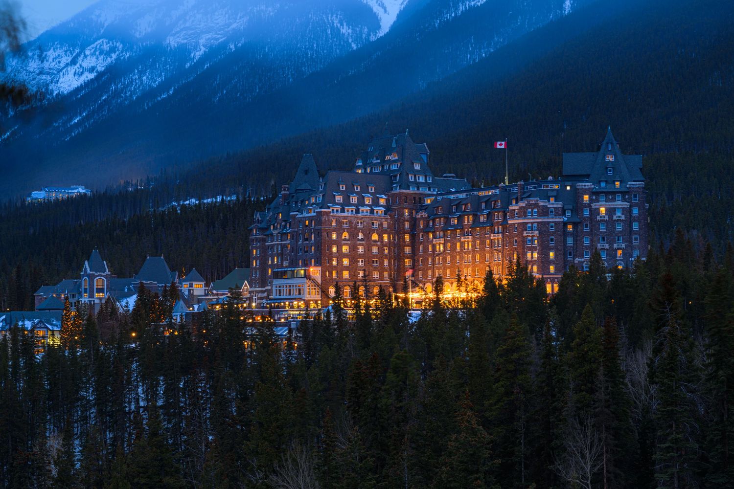 Illuminated Fairmont Banff Springs Hotel glowing at dusk surrounded by evergreen forest and misty mountain peaks in Canada.