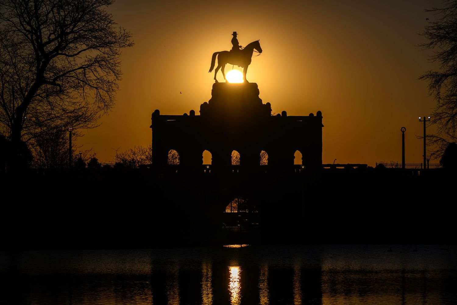 Silhouette of an equestrian statue atop a monument glowing against a golden sunset sky reflected in water below.