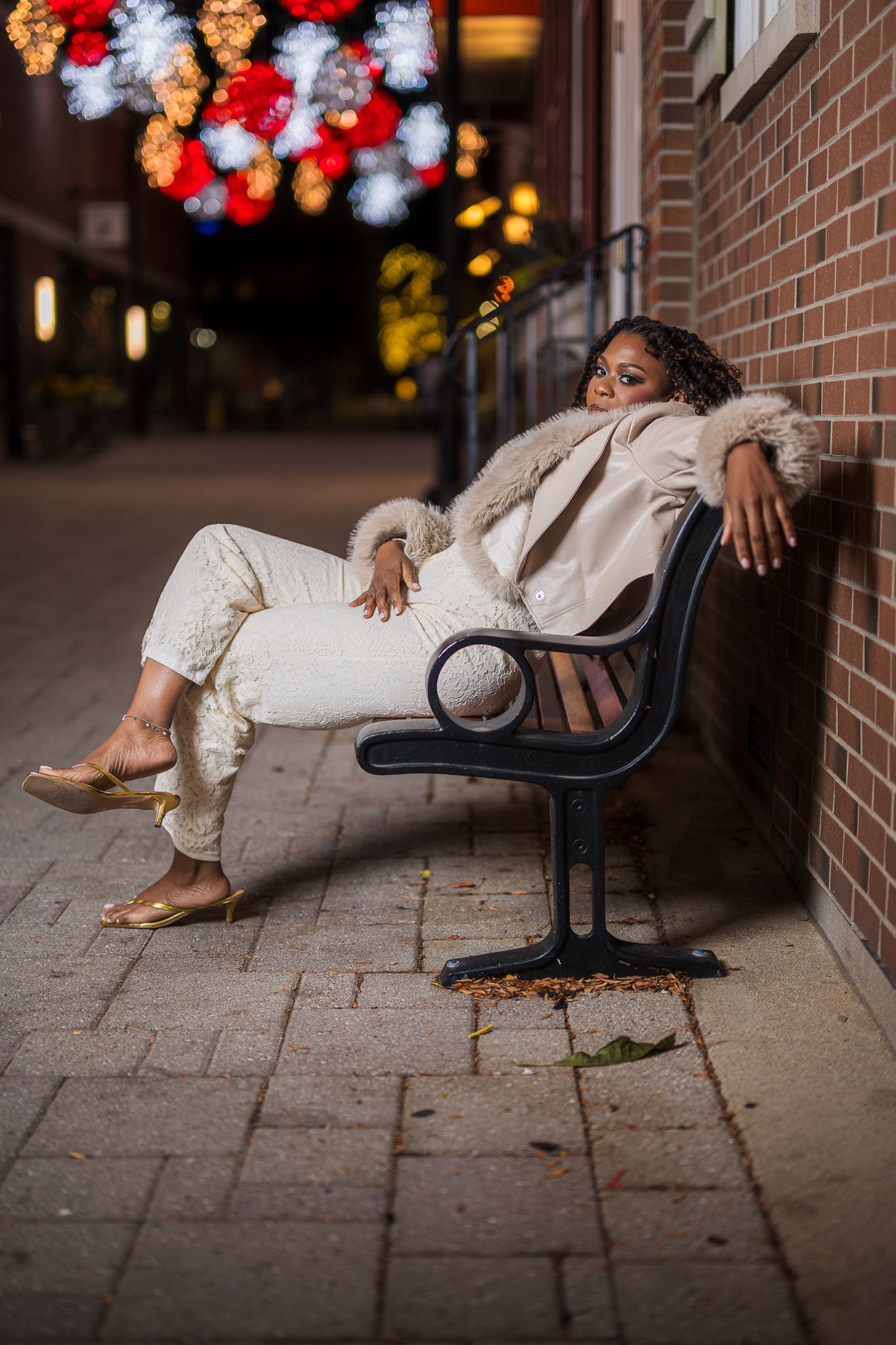 A stylish woman in a cream fur coat and white pants reclines on a bench against a brick wall with colorful bokeh lights.