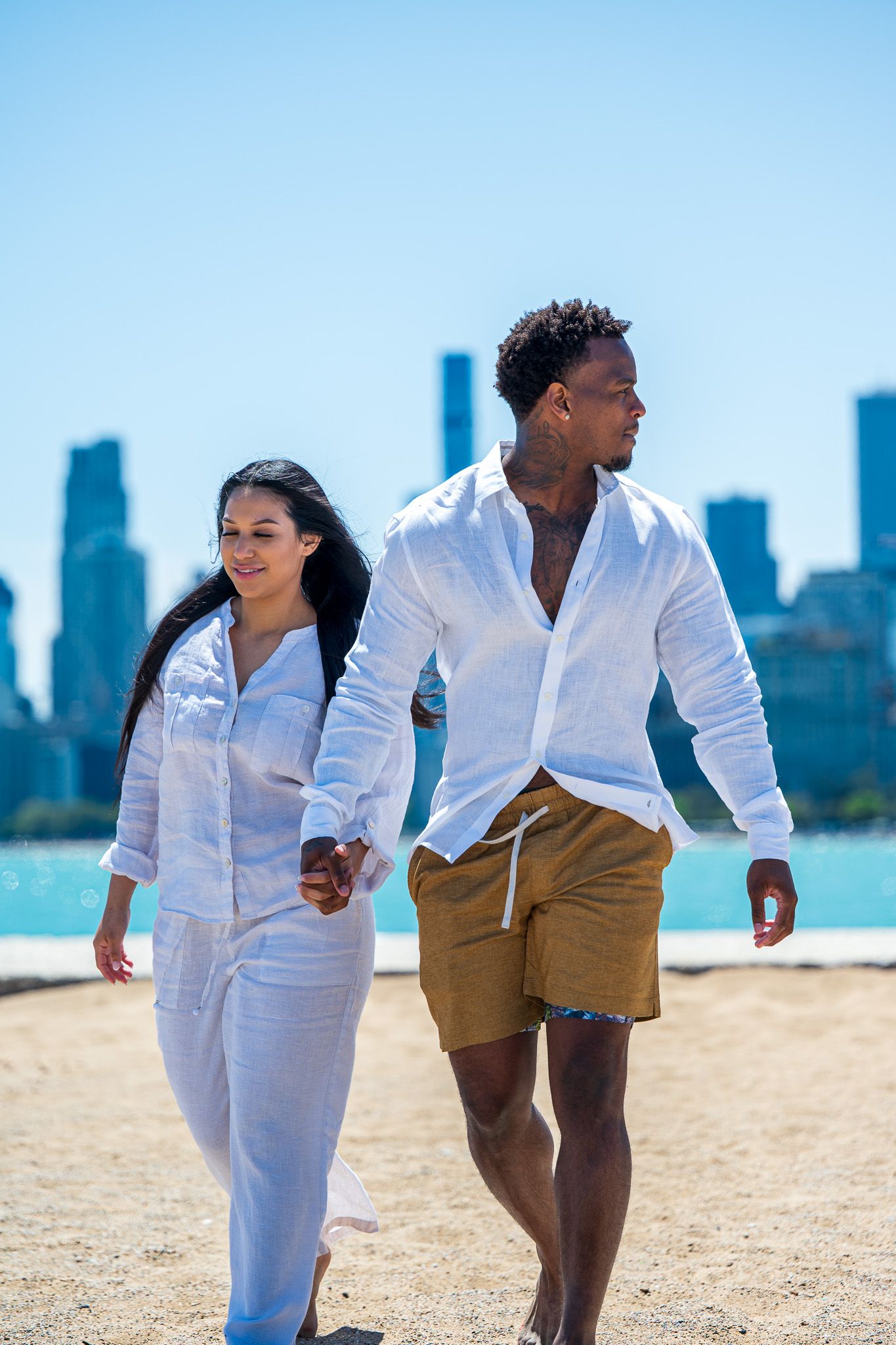 Interracial couple holding hands walking on sandy beach with city skyline in background on sunny day.