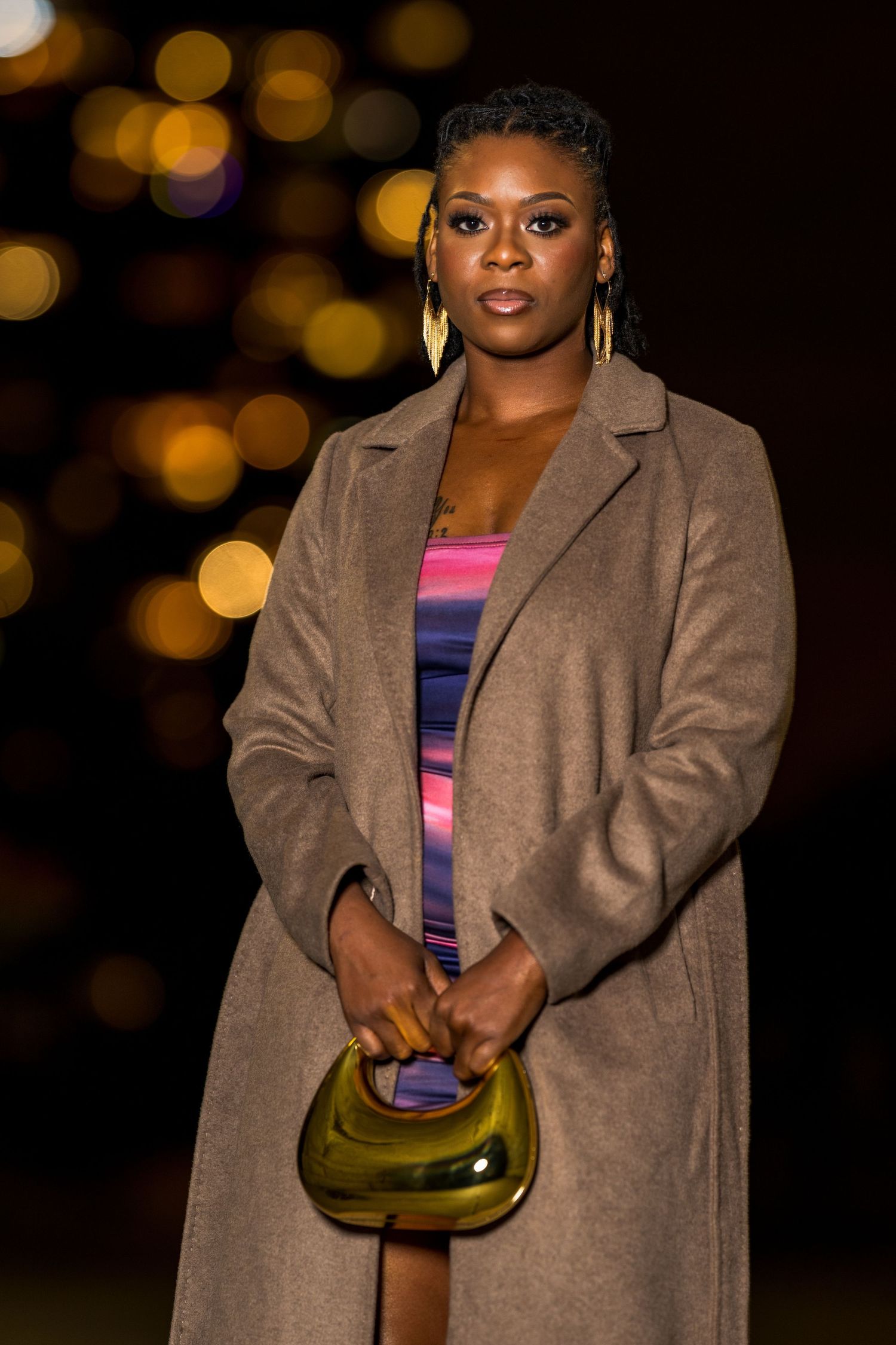 Woman in tan coat and colorful dress holds gold clutch purse outdoors at night with bokeh lights.