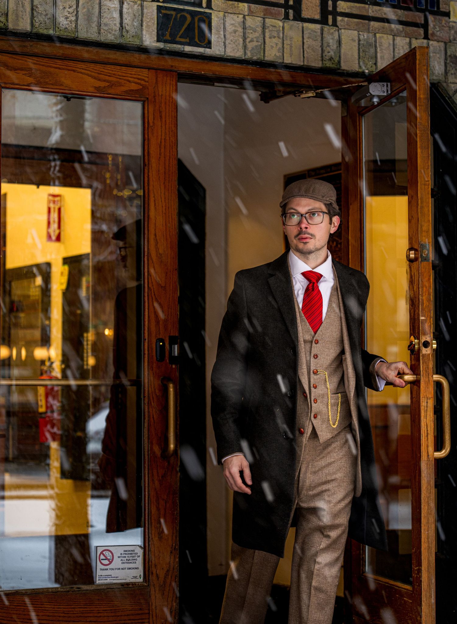 Man in vintage suit, red tie, and flat cap exits a building into falling snow through wooden doors.