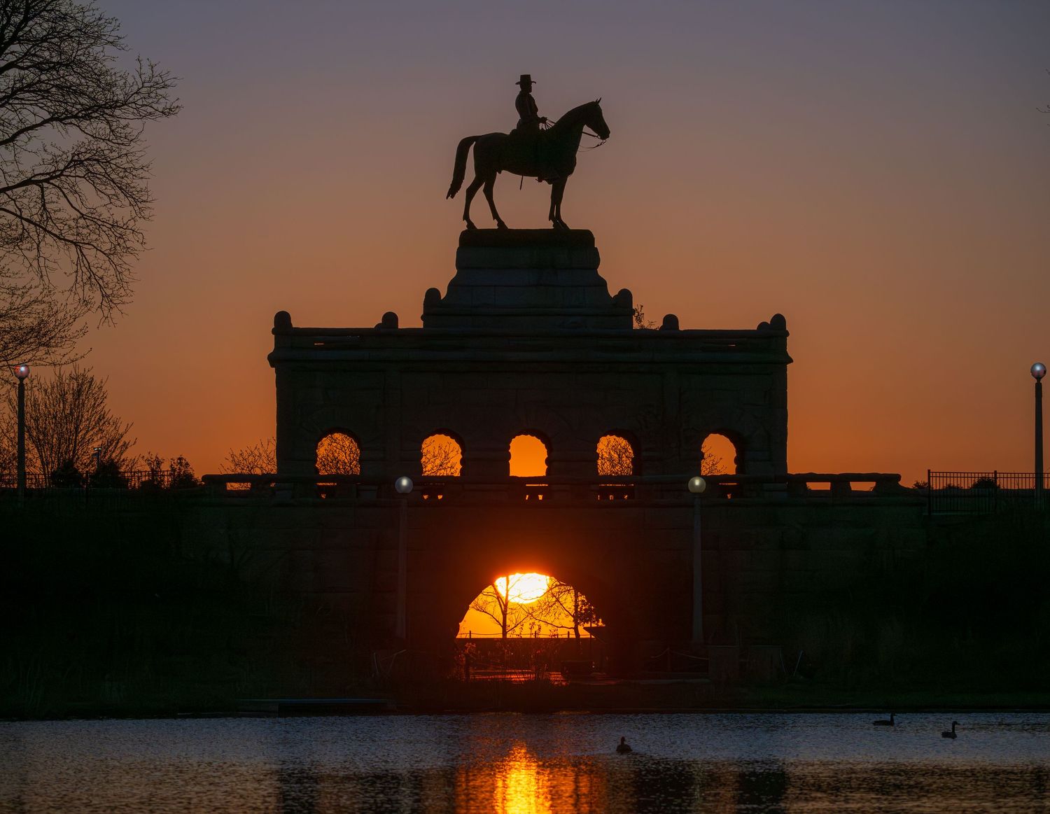 Silhouette of equestrian statue atop arched monument reflected in lake at golden sunset.